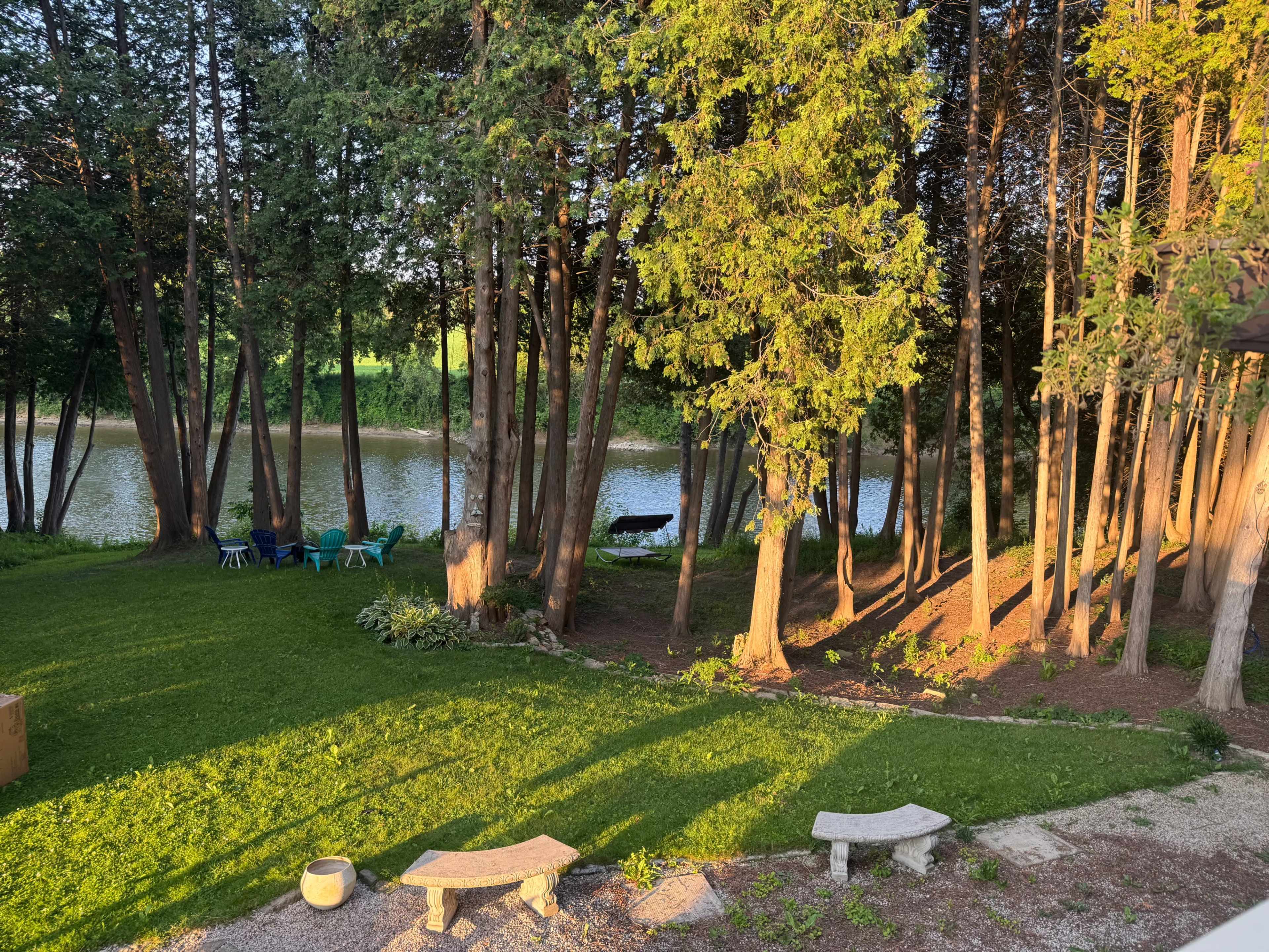 The image shows a tranquil riverside scene with green grass, trees, and a few chairs facing the water.