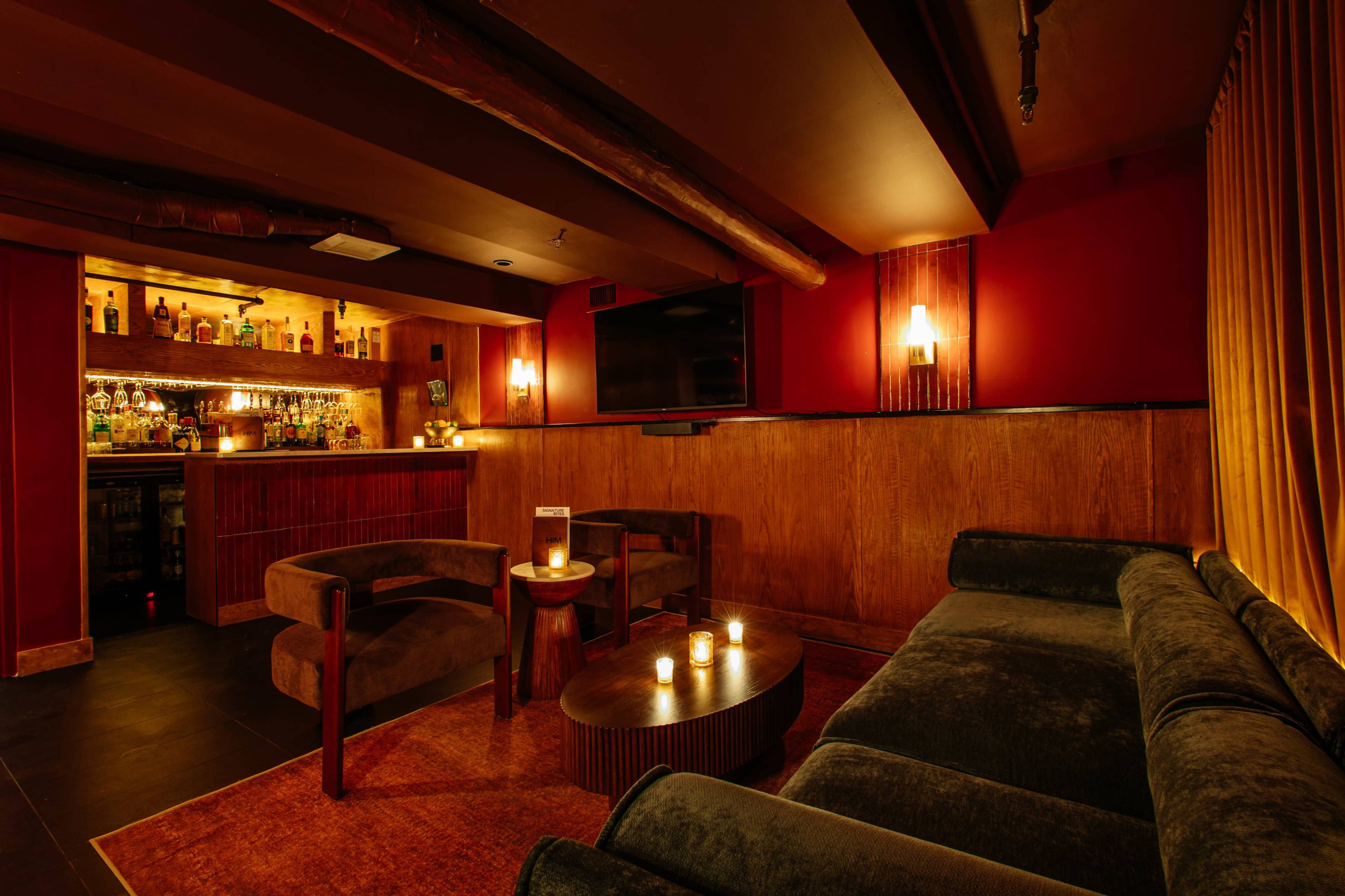 The image shows a dimly lit lounge with a bar in the background, featuring a seating area with dark chairs and a table adorned with candles.