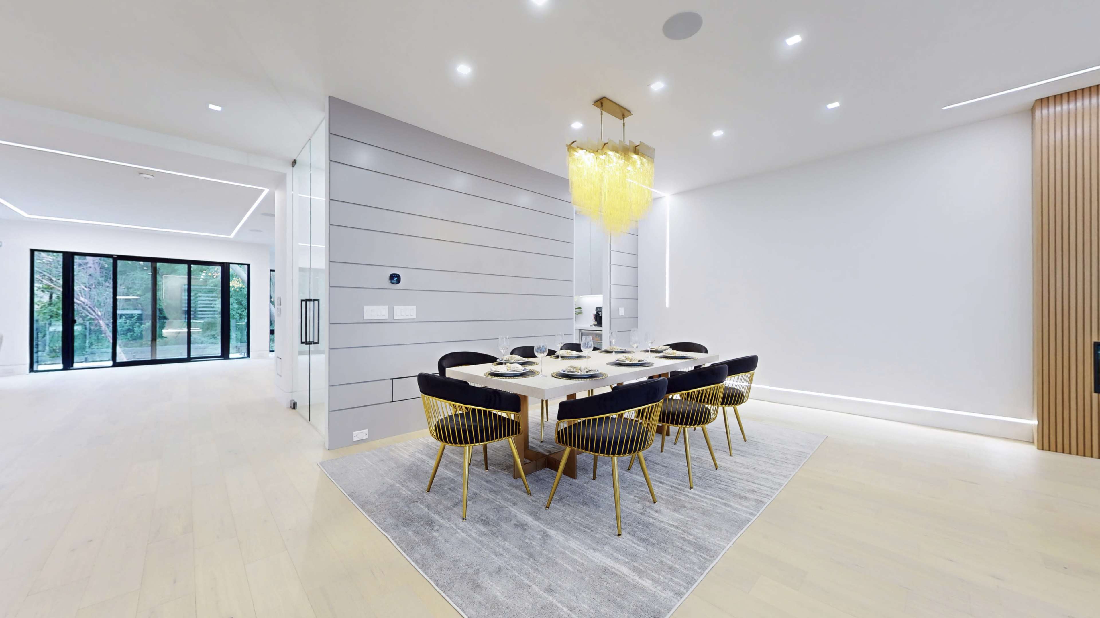 A modern dining area features a table set for six, surrounded by black and gold chairs, with a large light fixture above and a large glass door leading outside.