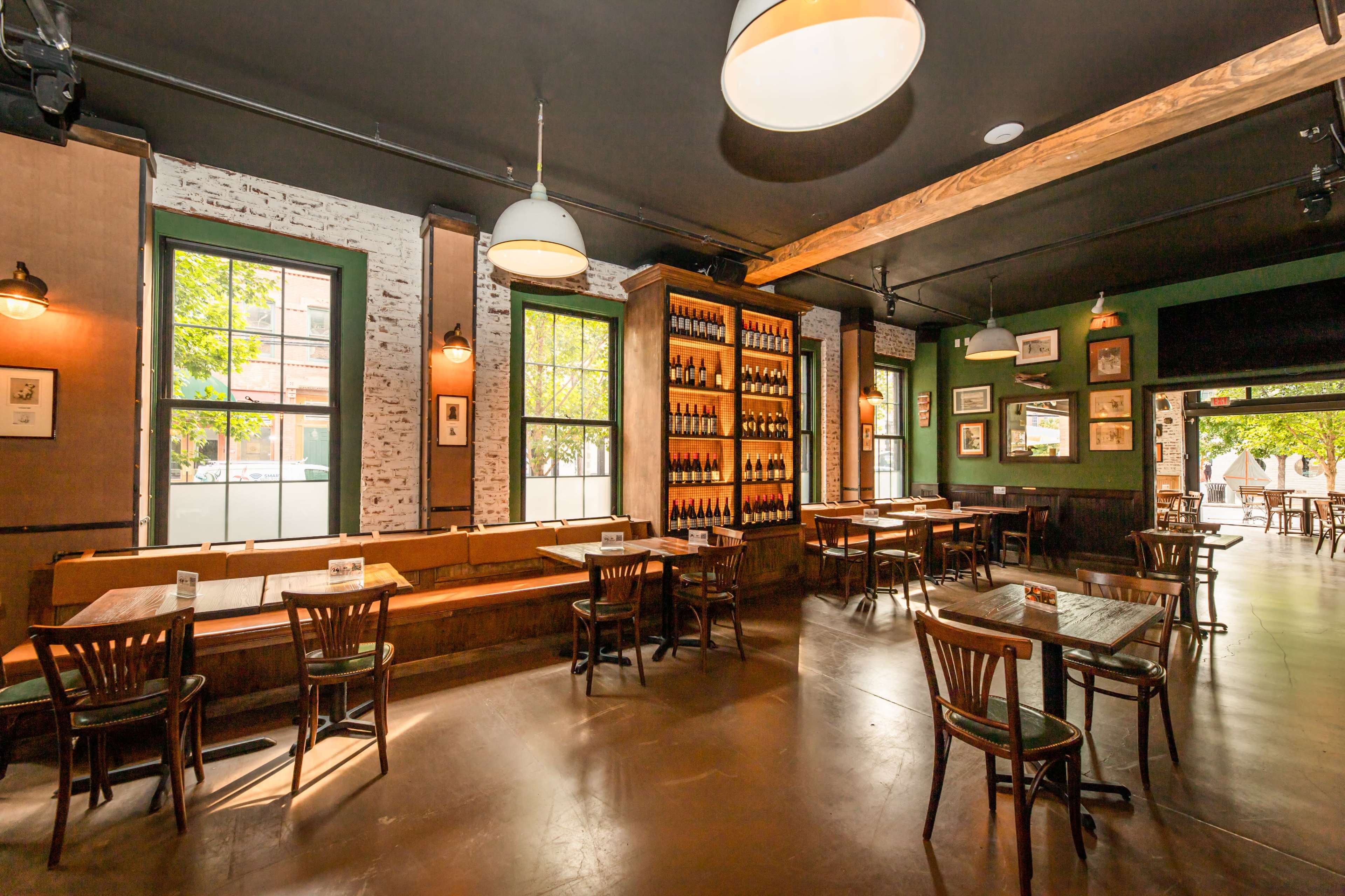 An interior of a restaurant featuring wooden tables, chairs, and a wall lined with shelves of bottles, illuminated by pendant lights.