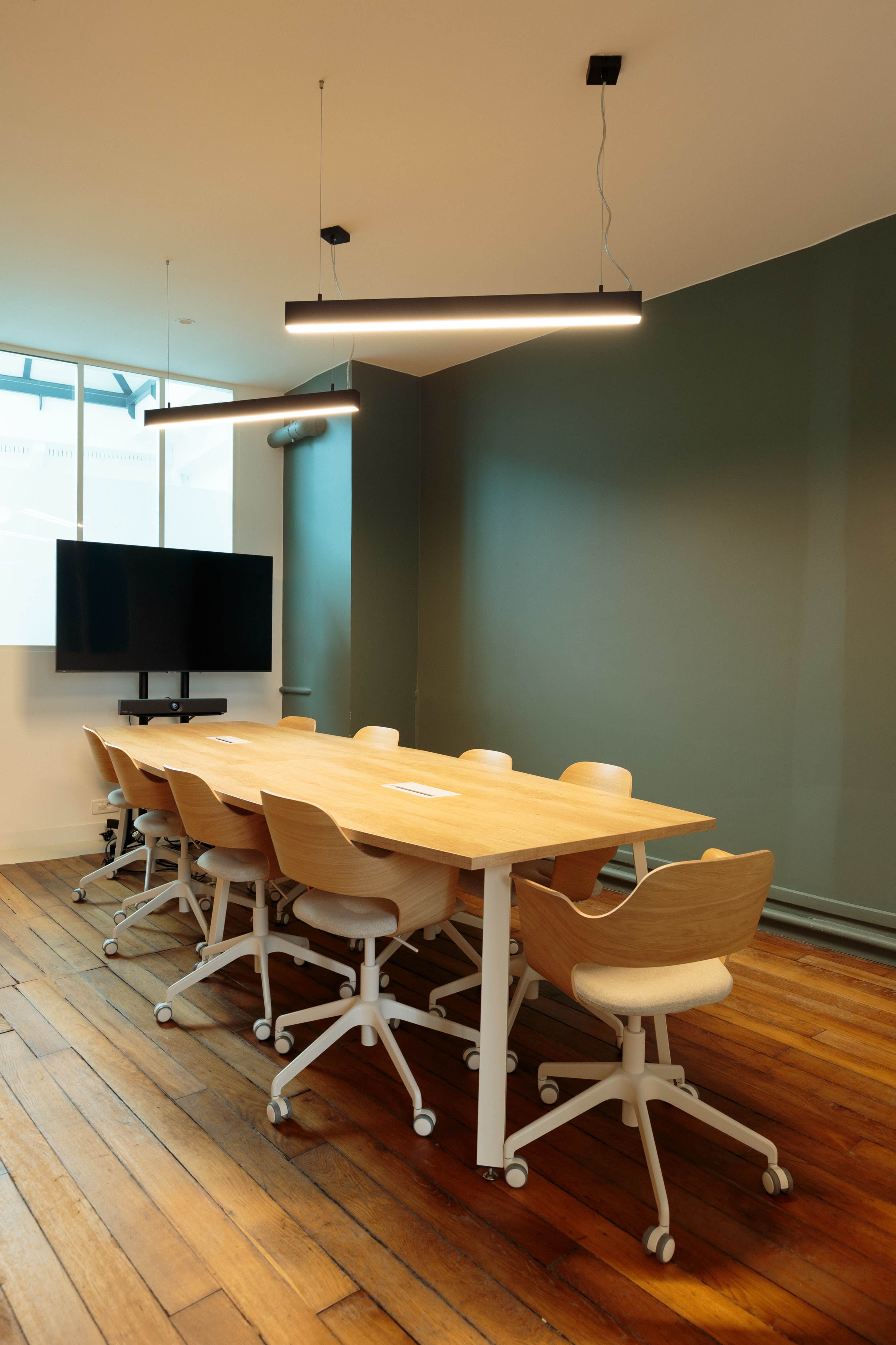 A modern conference room features a long wooden table surrounded by ergonomic chairs, with a wall-mounted TV and overhead lighting.