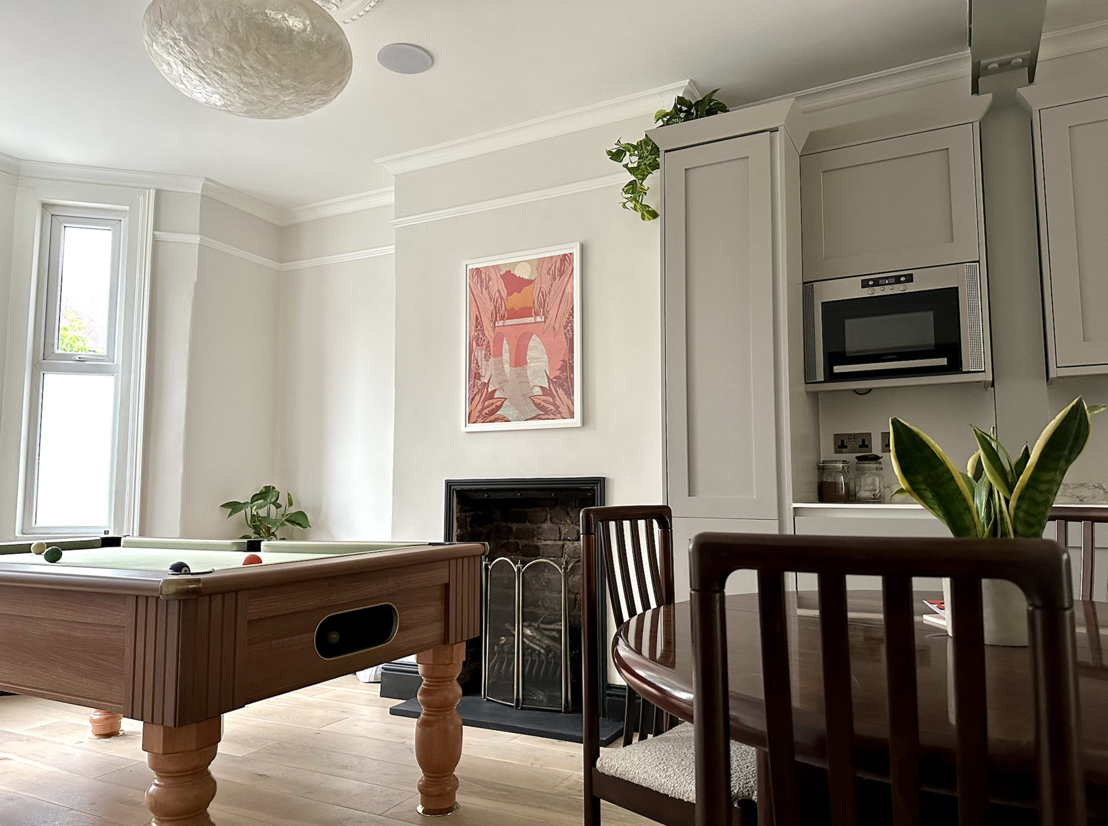 A modern living area features a billiard table, a dining table, and a framed artwork on the wall, alongside a fireplace and a microwave built into the cabinetry.
