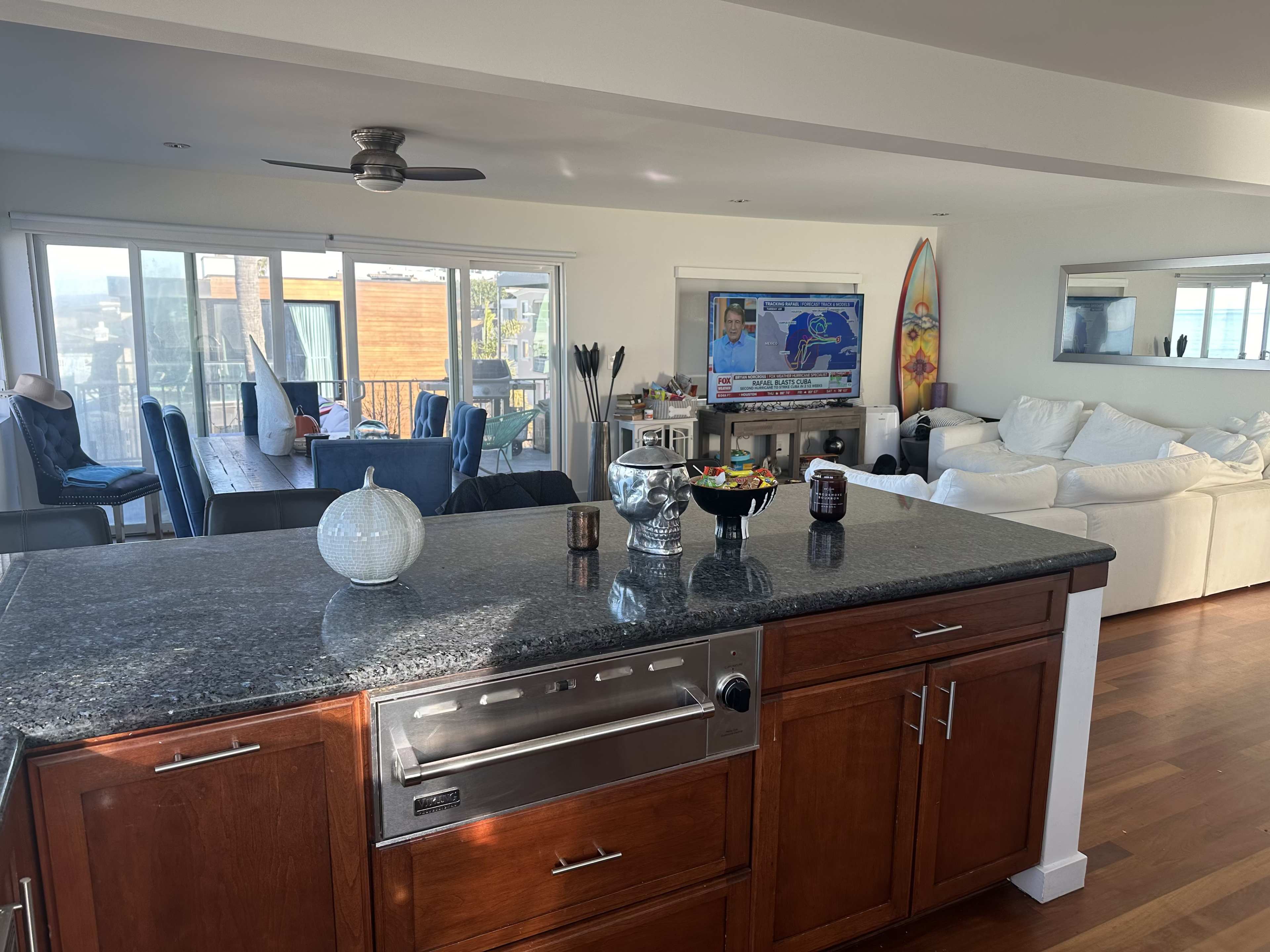 The image shows a modern kitchen with dark granite countertops, wooden cabinets, and an open layout leading to a living area with white sofas and a large TV displaying a weather report.