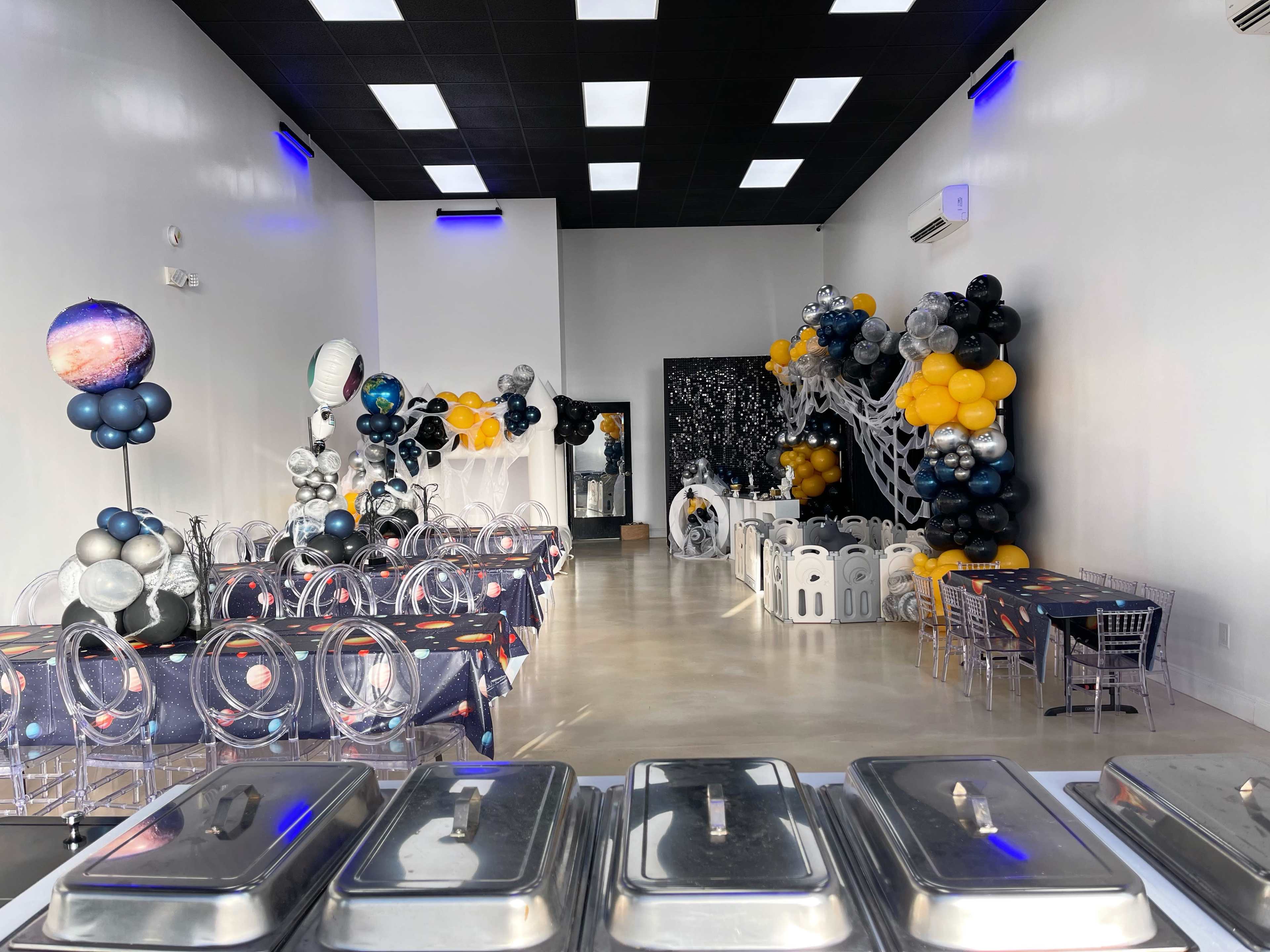 A spacious event hall decorated with space-themed balloons, tables covered with cosmic tablecloths, and a buffet setup in the foreground.