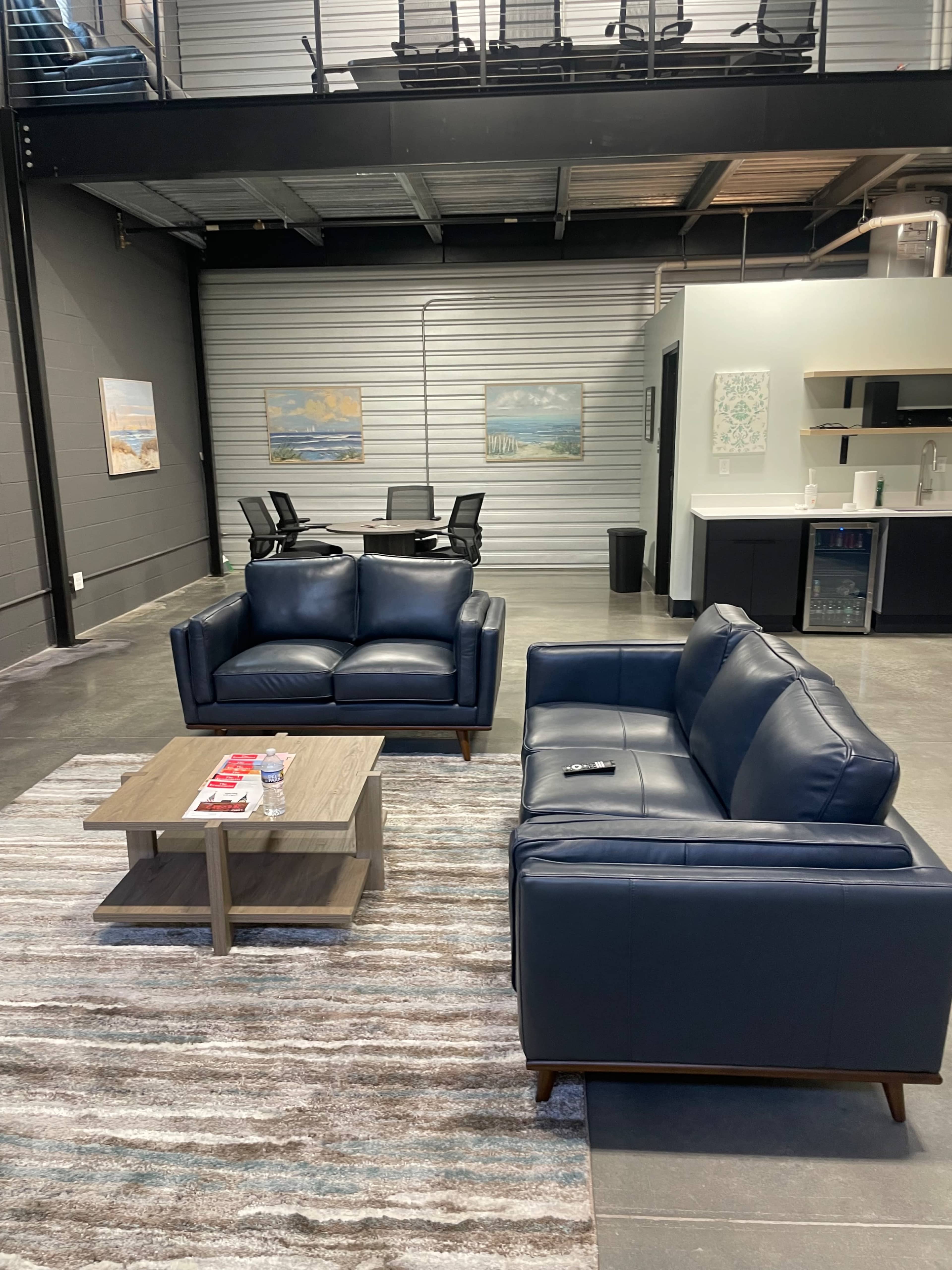 The image shows a modern lounge area with two blue leather sofas facing a wooden coffee table, set on a striped area rug, and a kitchenette visible in the background.