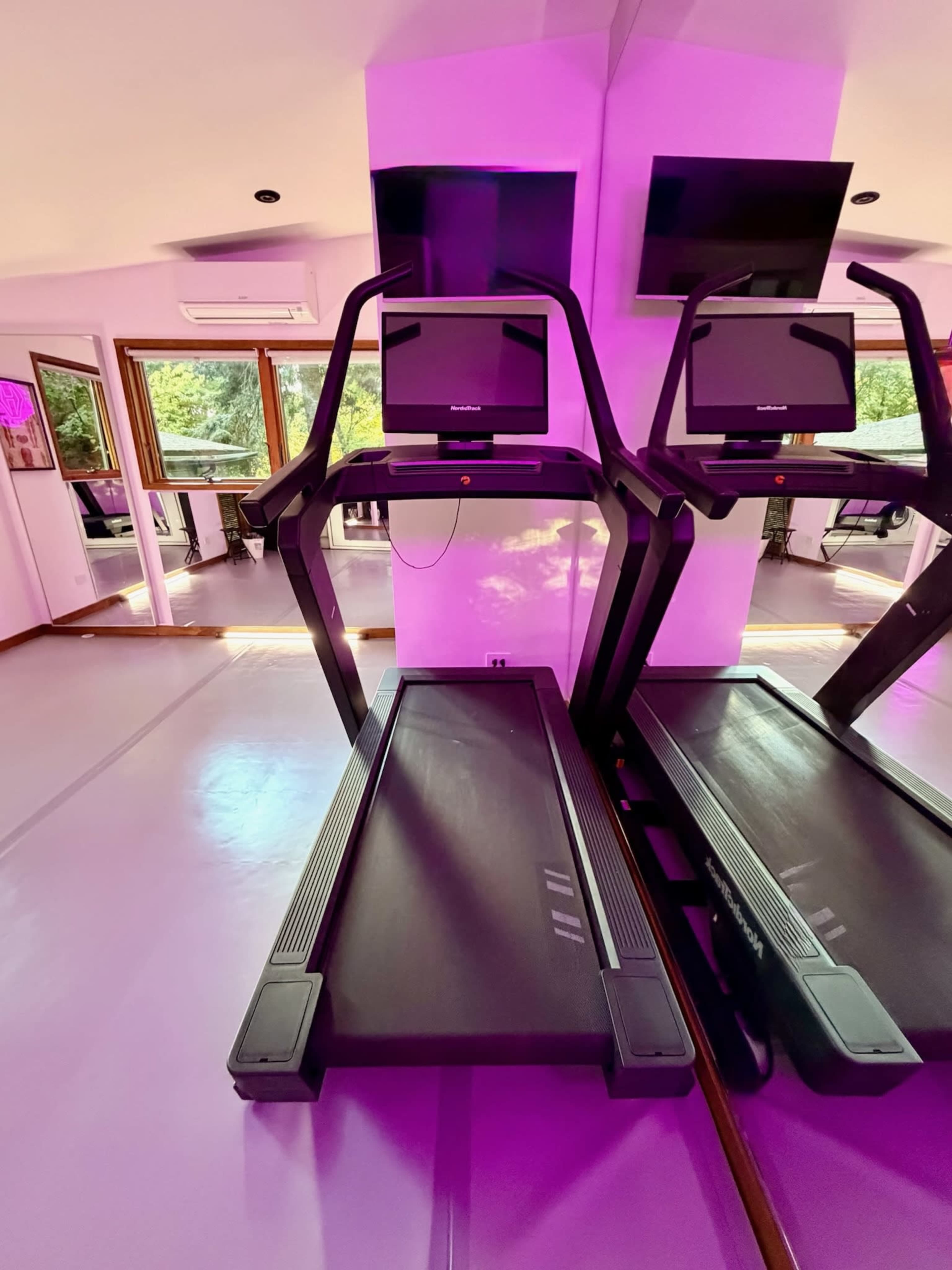 The image shows two treadmills facing a mirrored wall in a brightly lit gym with purple accents.