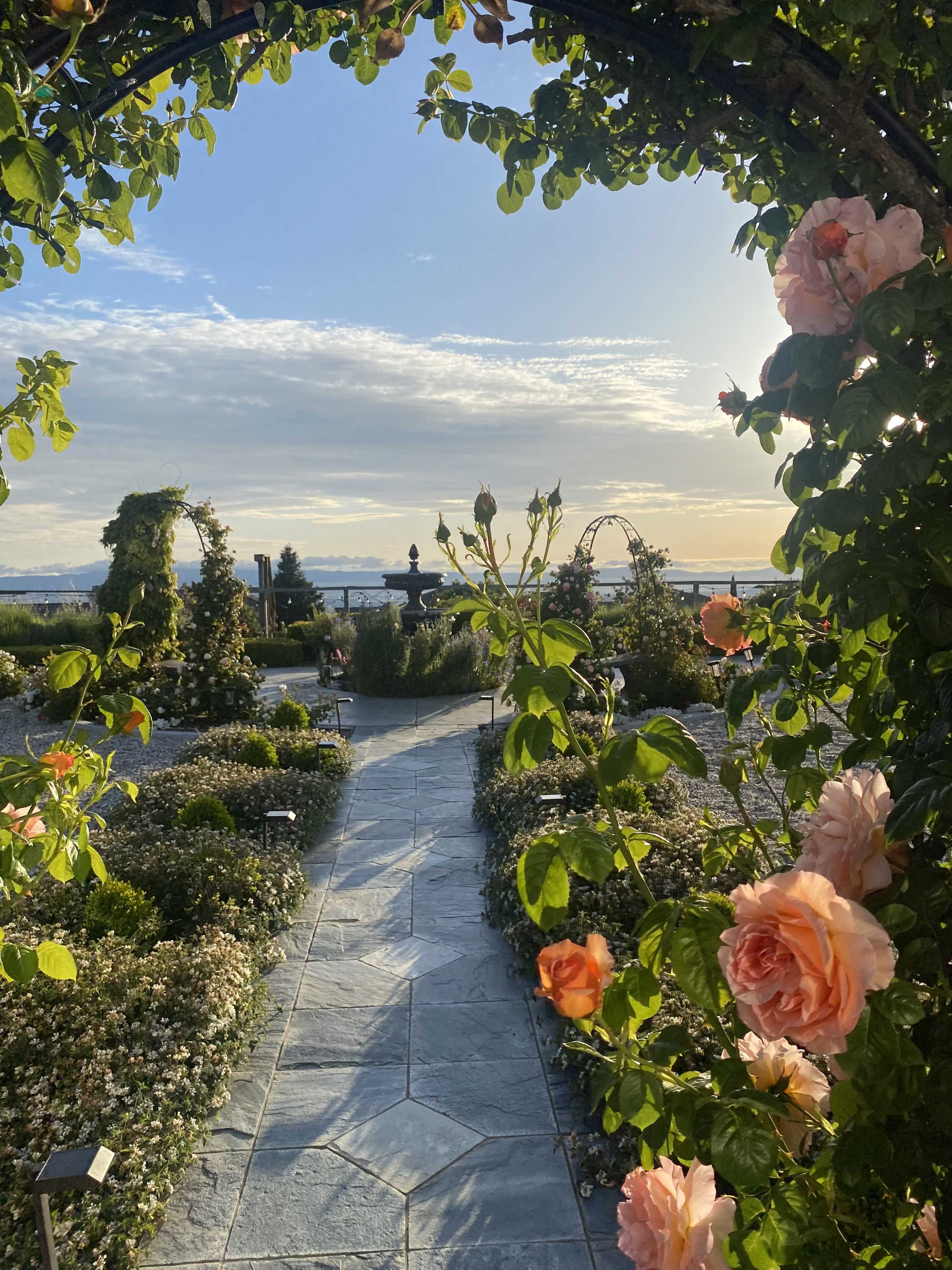 A stone pathway lined with flowers leads through an archway adorned with pink roses toward a garden and a distant skyline under a clear sky.
