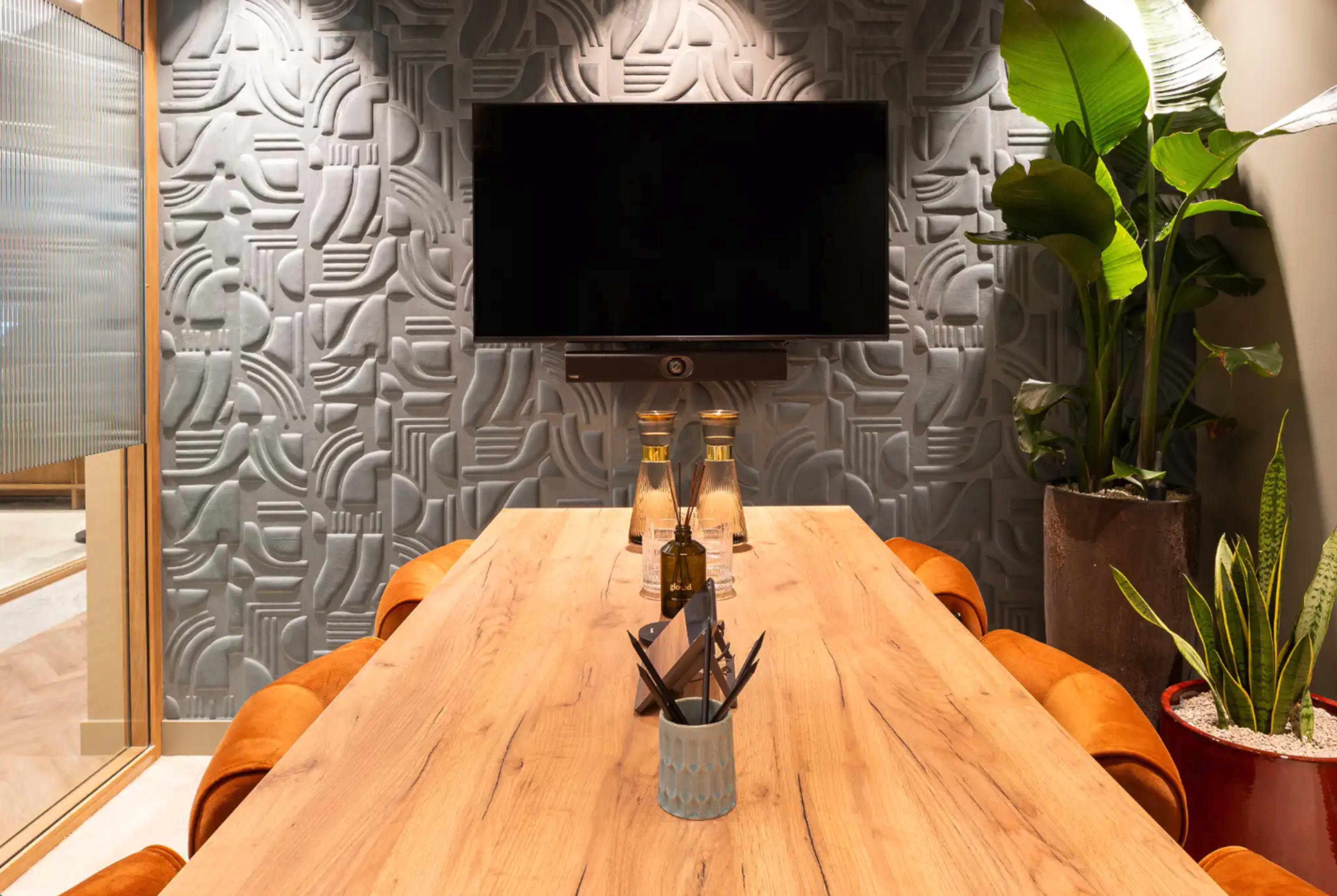 A modern conference room features a wooden table with leather chairs, a wall-mounted TV, and textured wallpaper, accompanied by indoor plants.