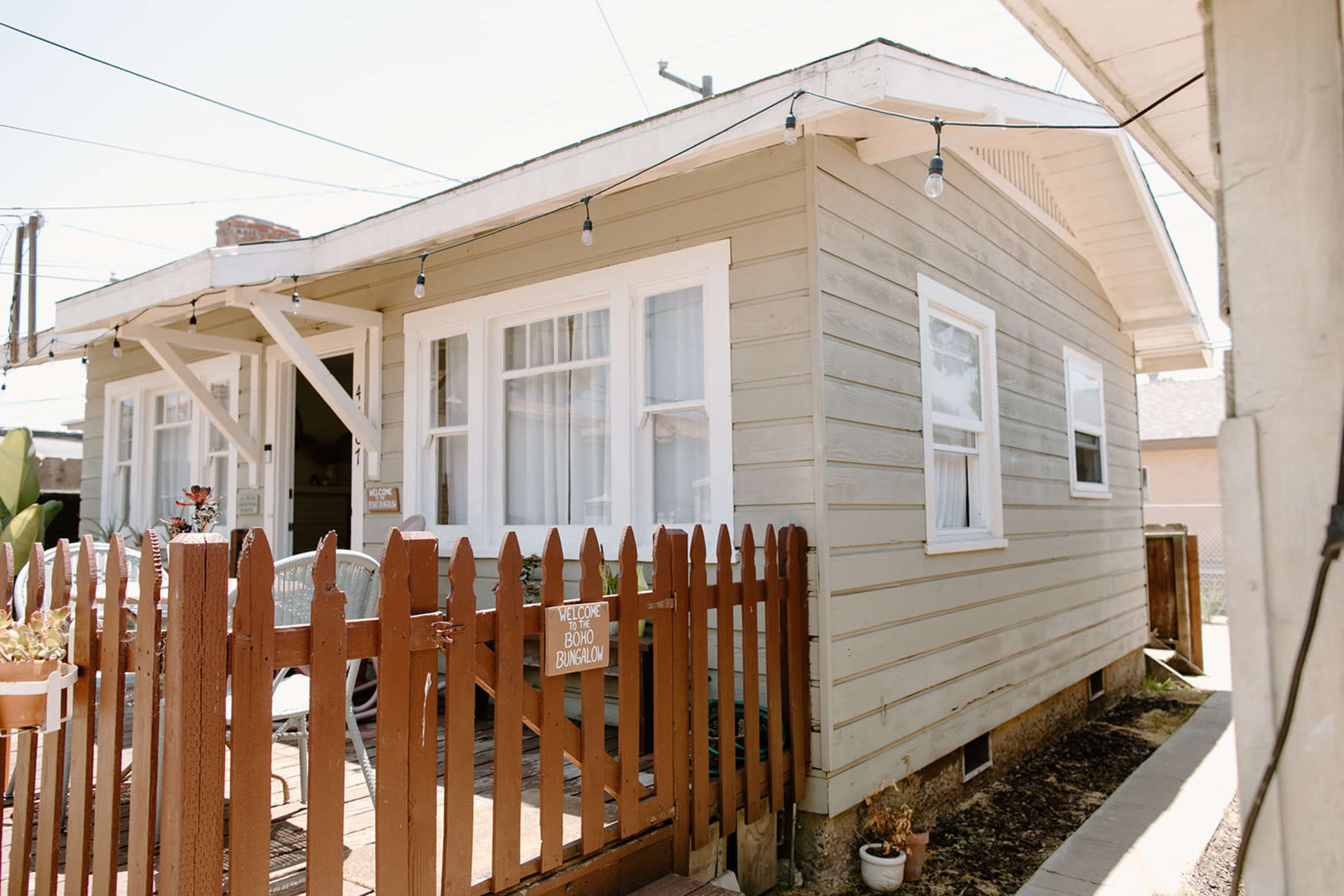 A single-story, light-colored wooden house with a front porch and a brown picket fence is shown.