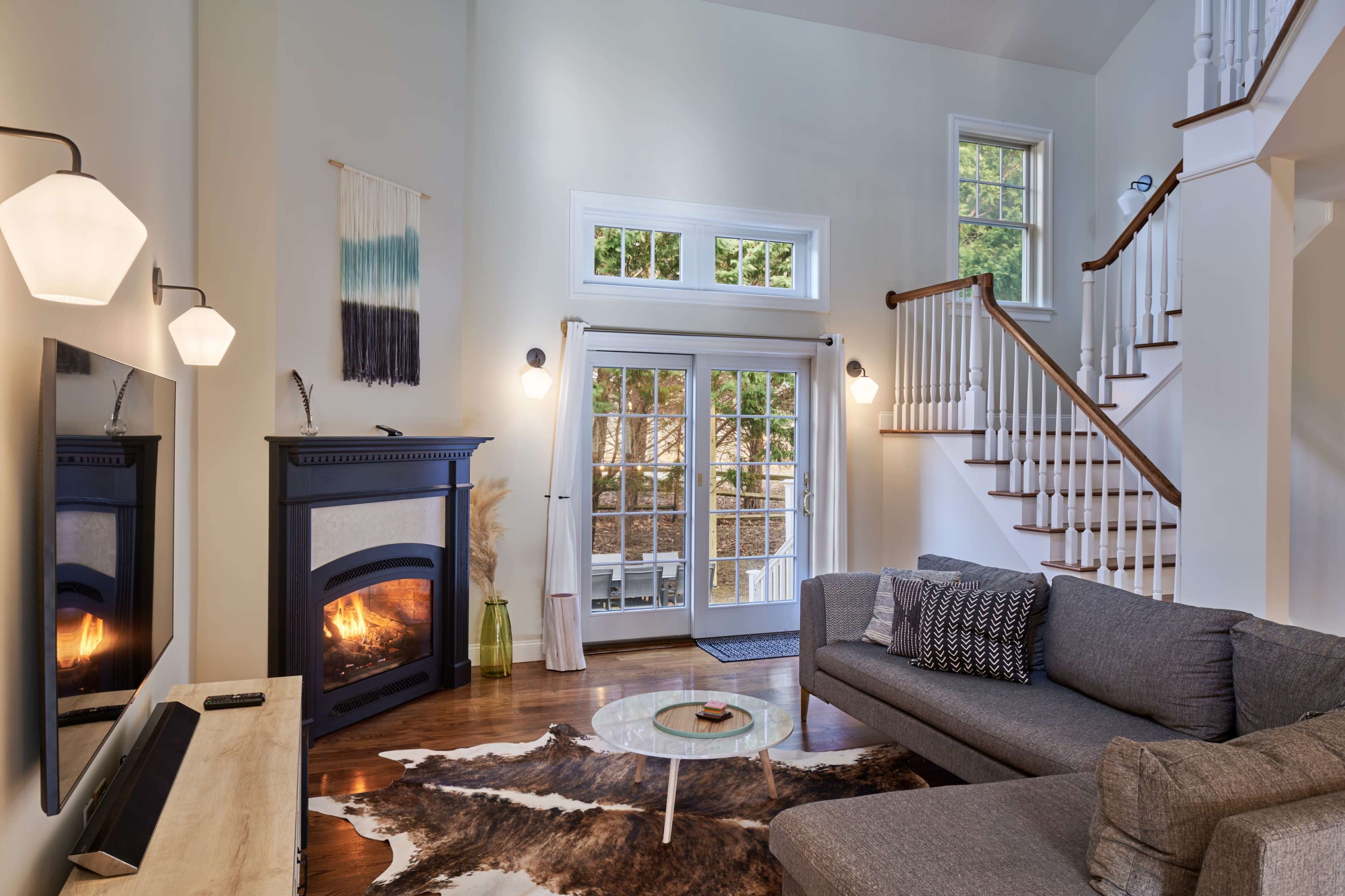 A spacious living room featuring a gray sectional sofa, a coffee table on a patterned rug, a fireplace, and large windows that allow natural light to fill the space.
