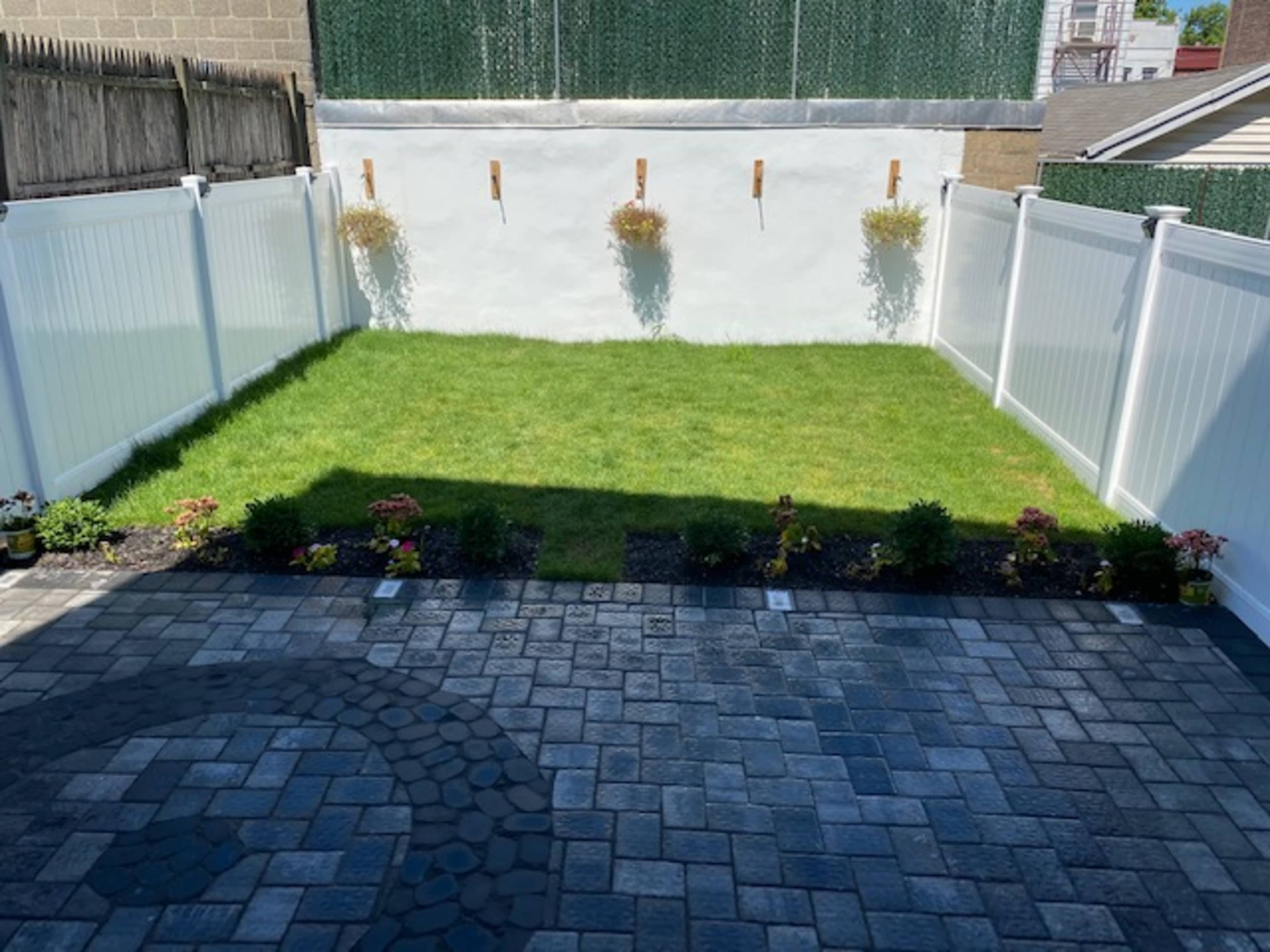 A small, fenced backyard with a grassy area surrounded by a stone patio and flower beds.