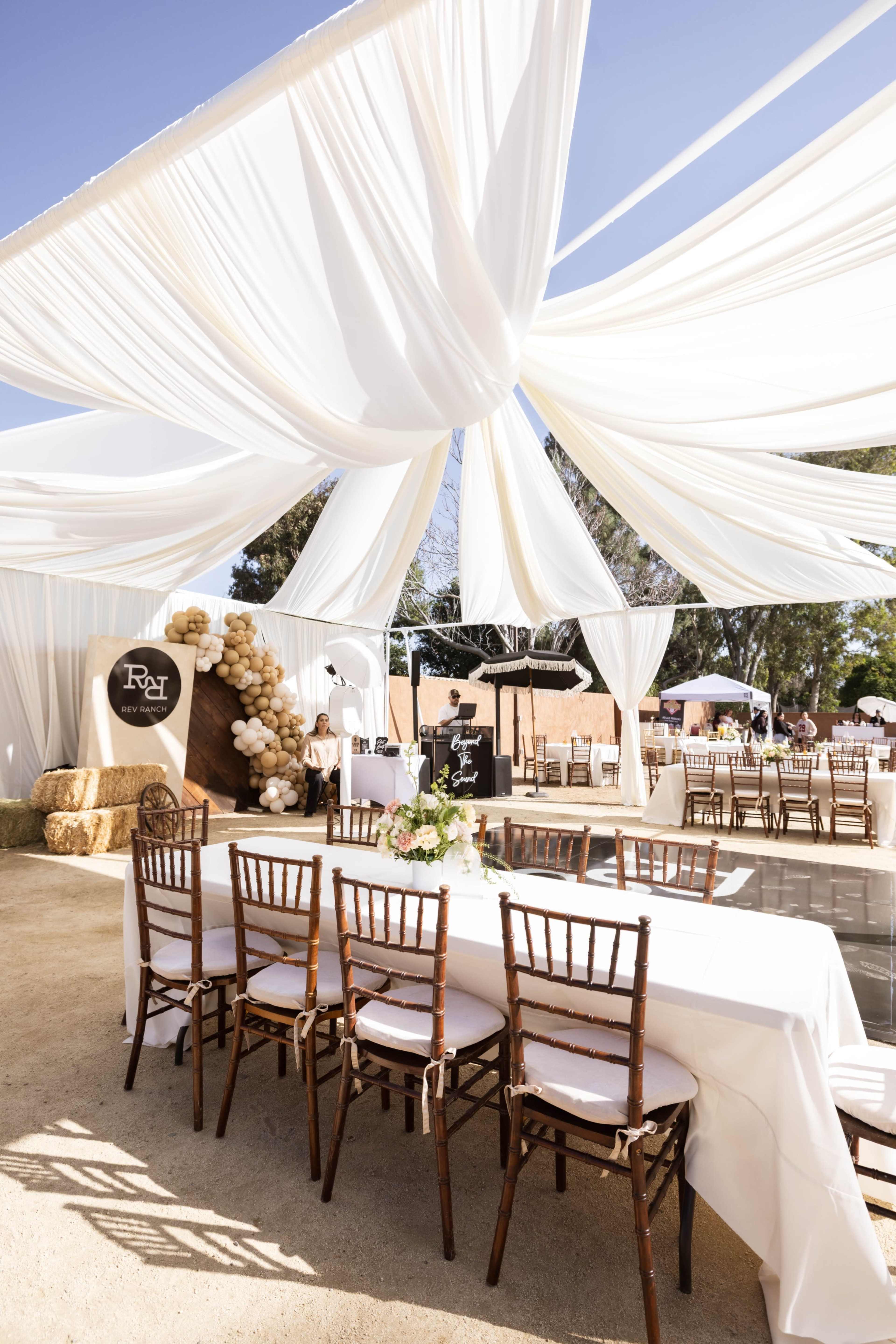 A large outdoor event space features elegant white drapery overhead, a long table set with chairs, and decorative elements, including a balloon arch and a backdrop.