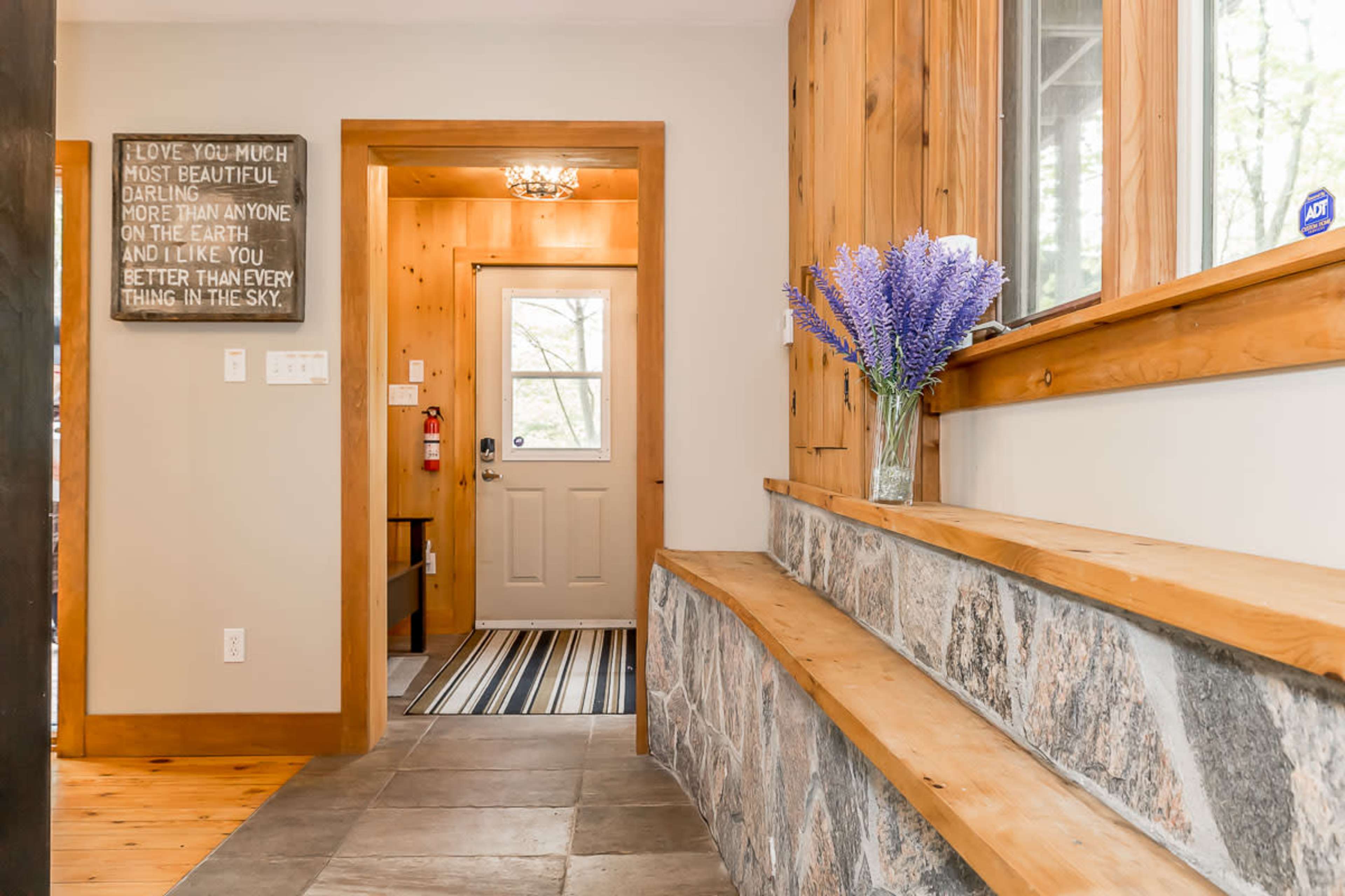 A cozy entryway with wooden walls, a stone bench, and a door leading outside, adorned with a vase of lavender flowers.