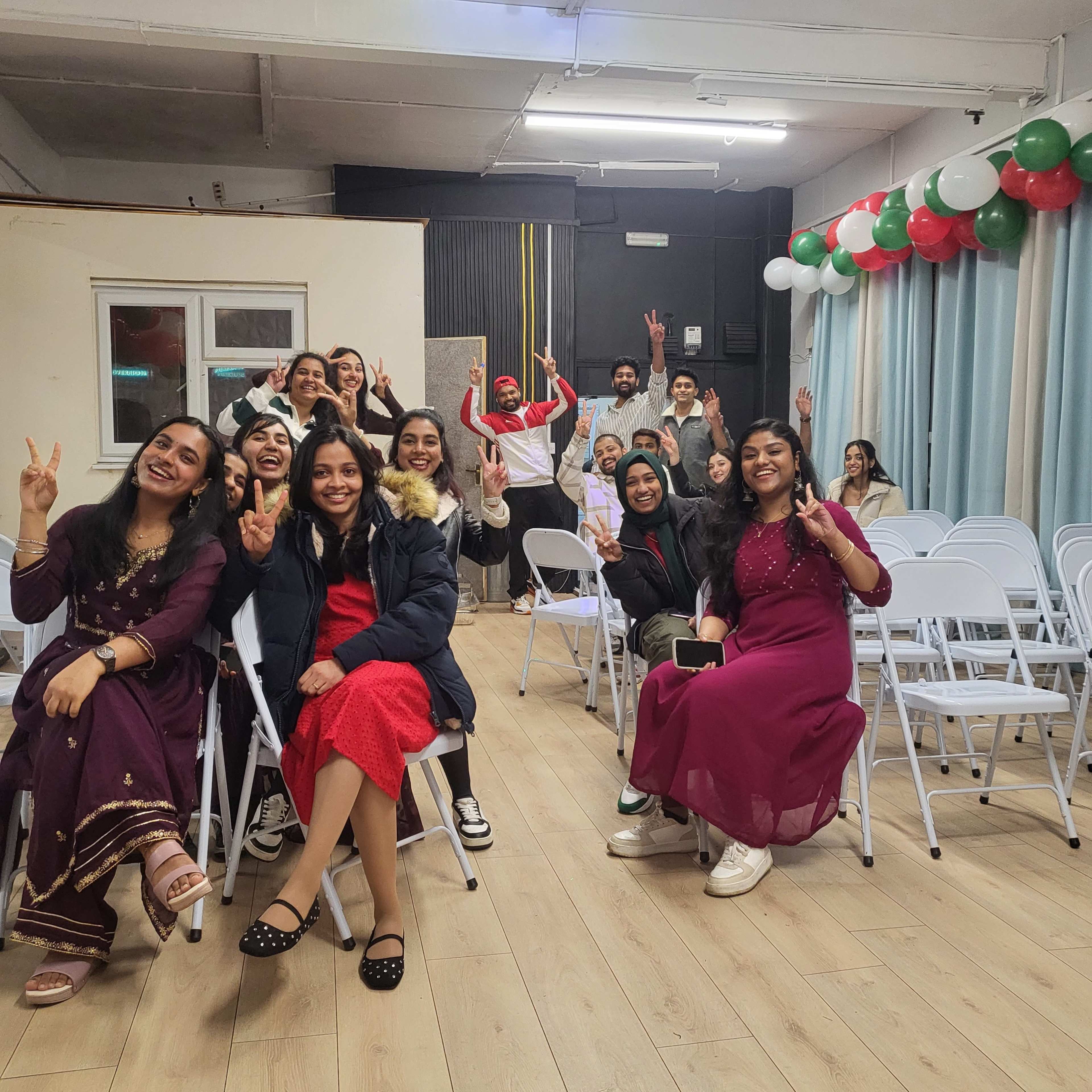 A group of people is seated in a room decorated with balloons, smiling and making peace signs.