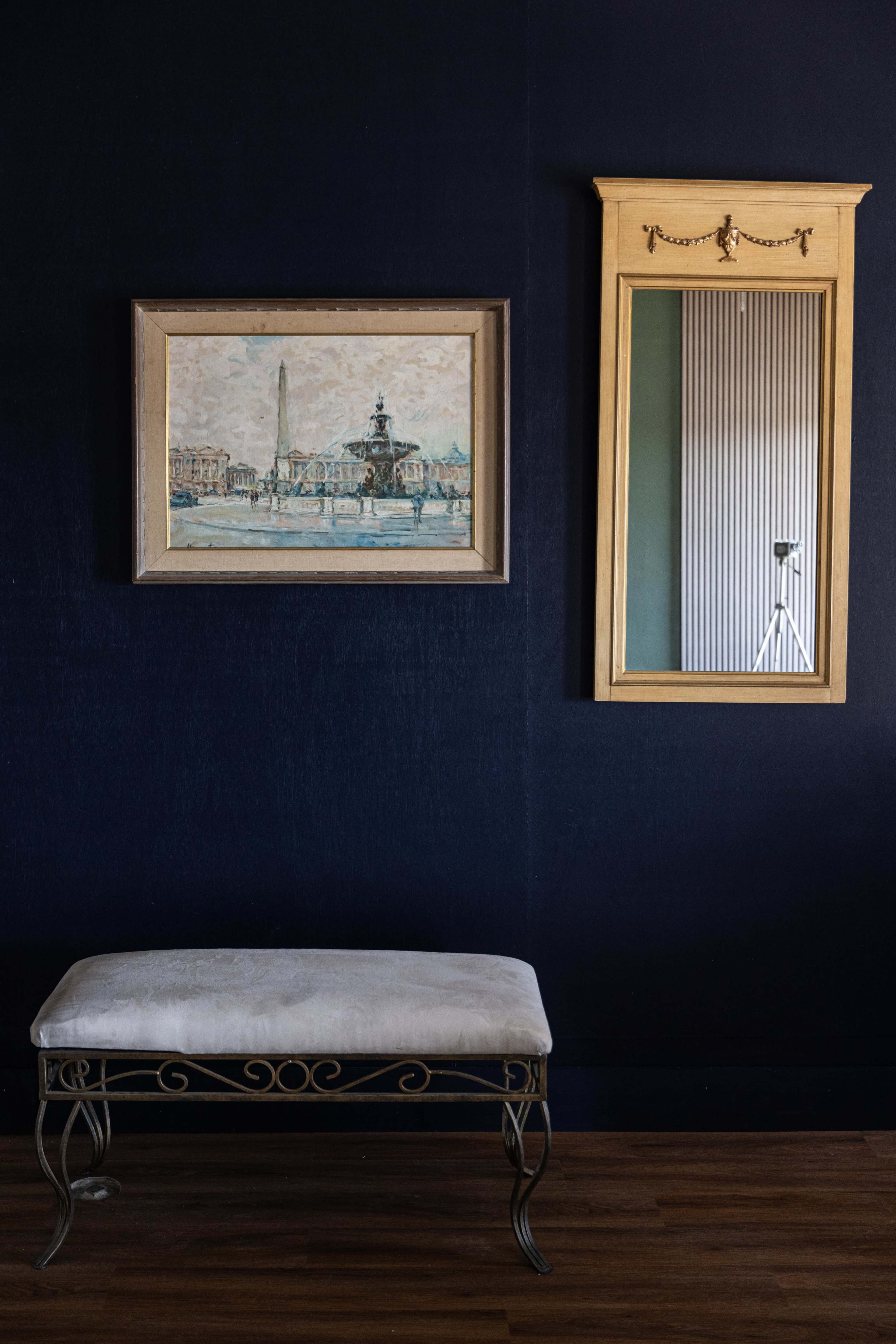 The image features a bench in front of a dark blue wall, with a framed painting on the left and a gold-framed mirror on the right.