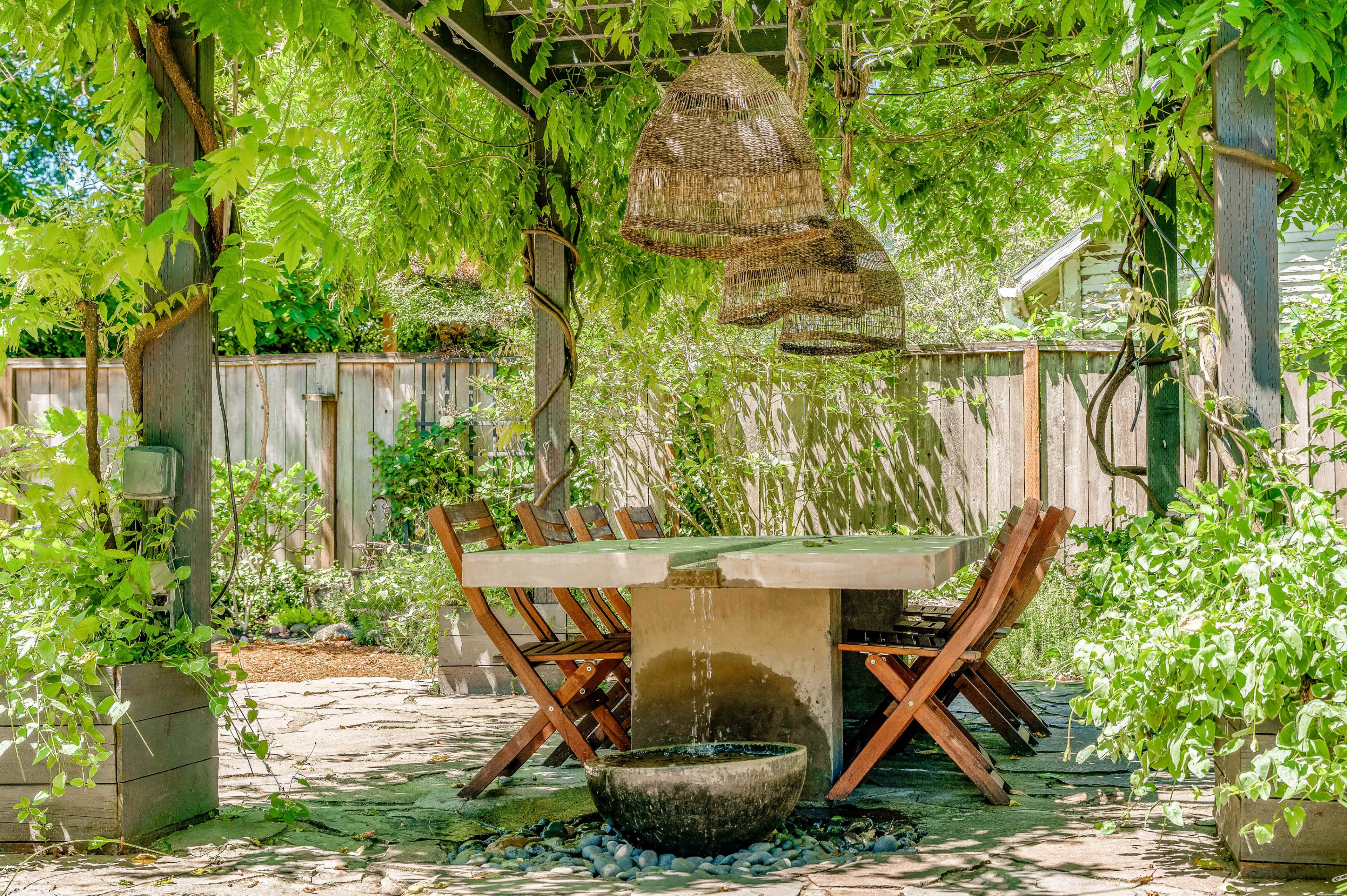 A shaded outdoor dining area features a large concrete table surrounded by wooden chairs and decorative hanging lights.