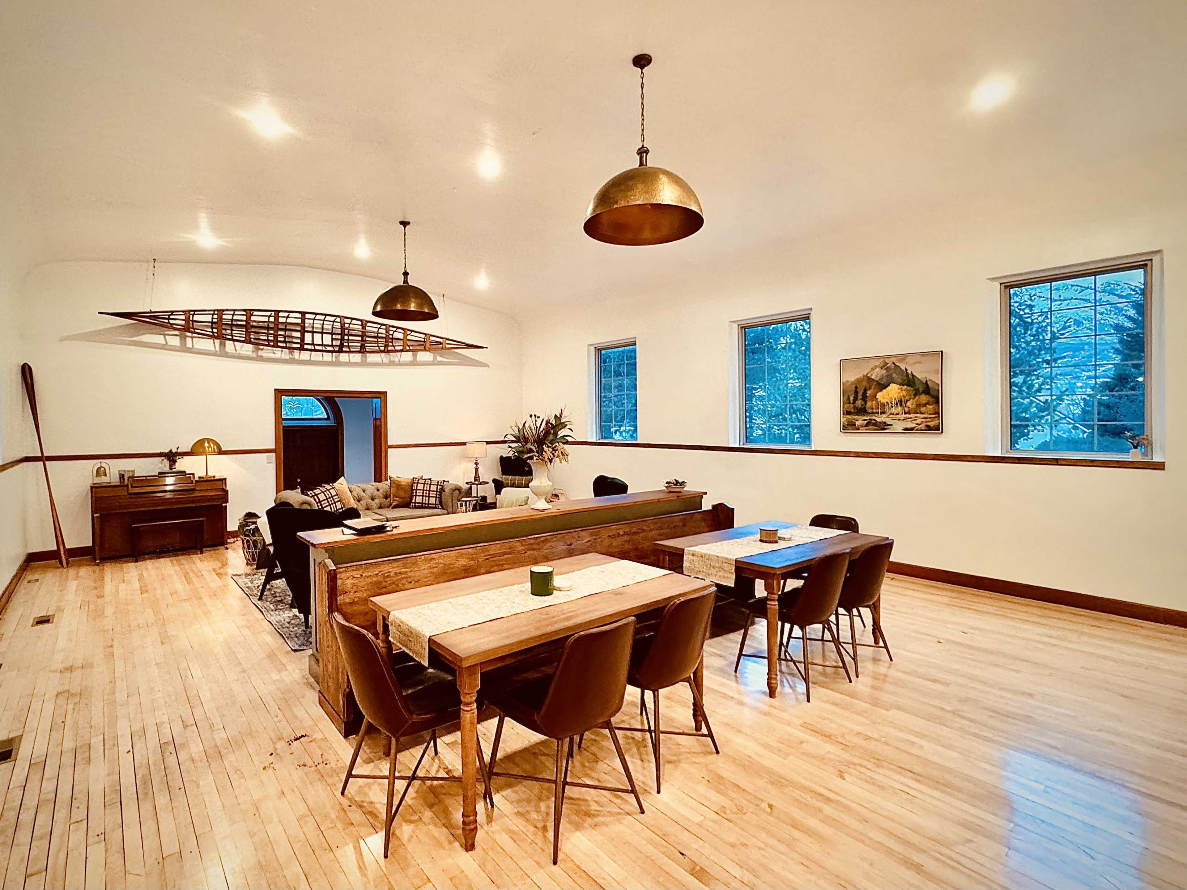 The image shows a spacious, well-lit dining and living area featuring a long wooden table, chairs, and decorative elements like a boat oar and artwork on the walls.
