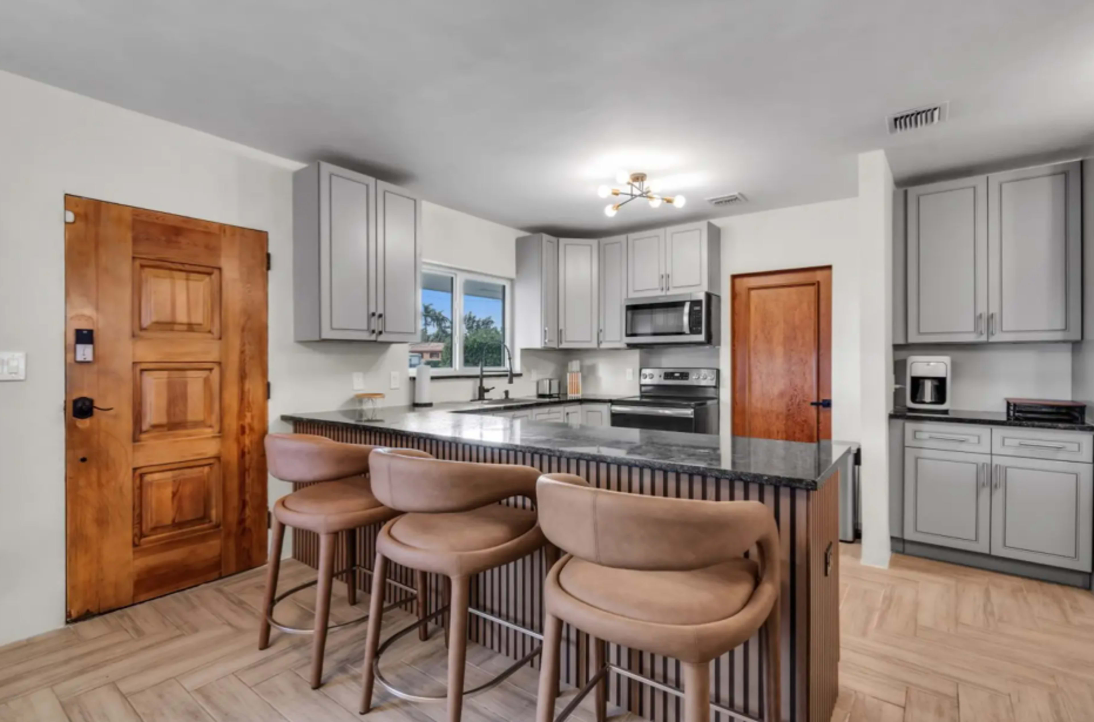 The modern kitchen features gray cabinets, a large island with bar stools, and stainless steel appliances.