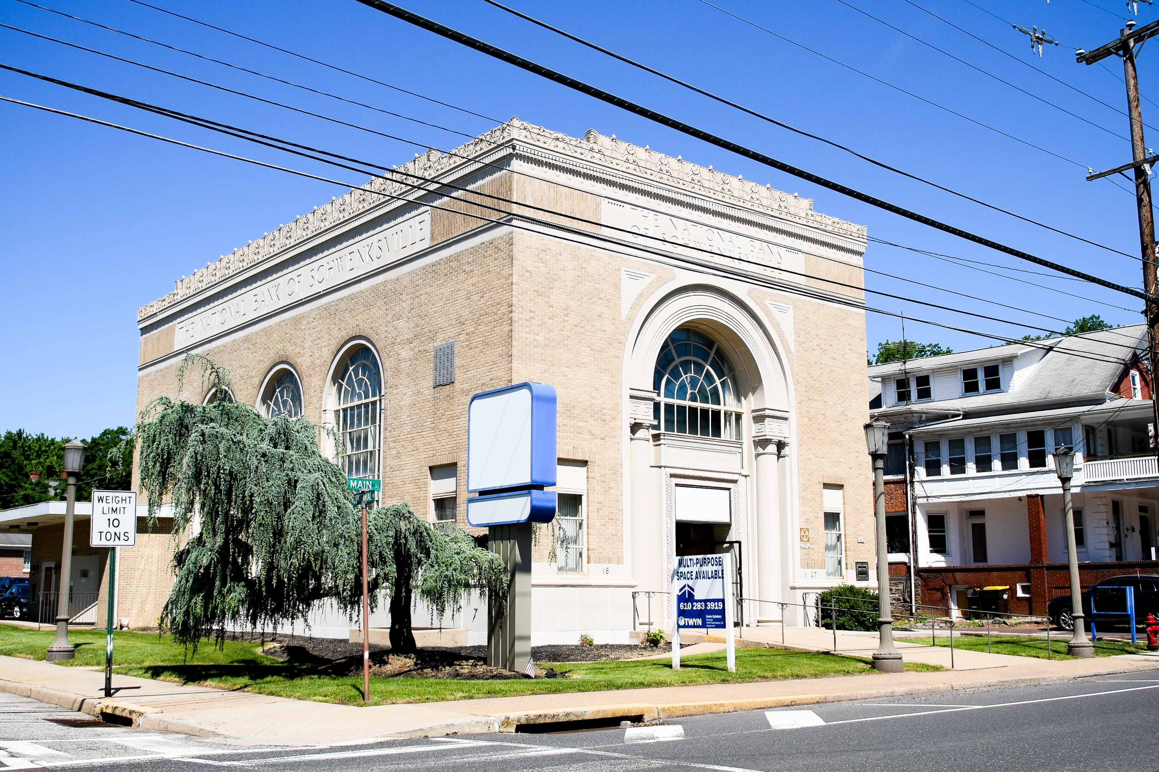 The image shows a historic building with large windows and decorative architectural elements, situated at an intersection near a tree and a street sign.