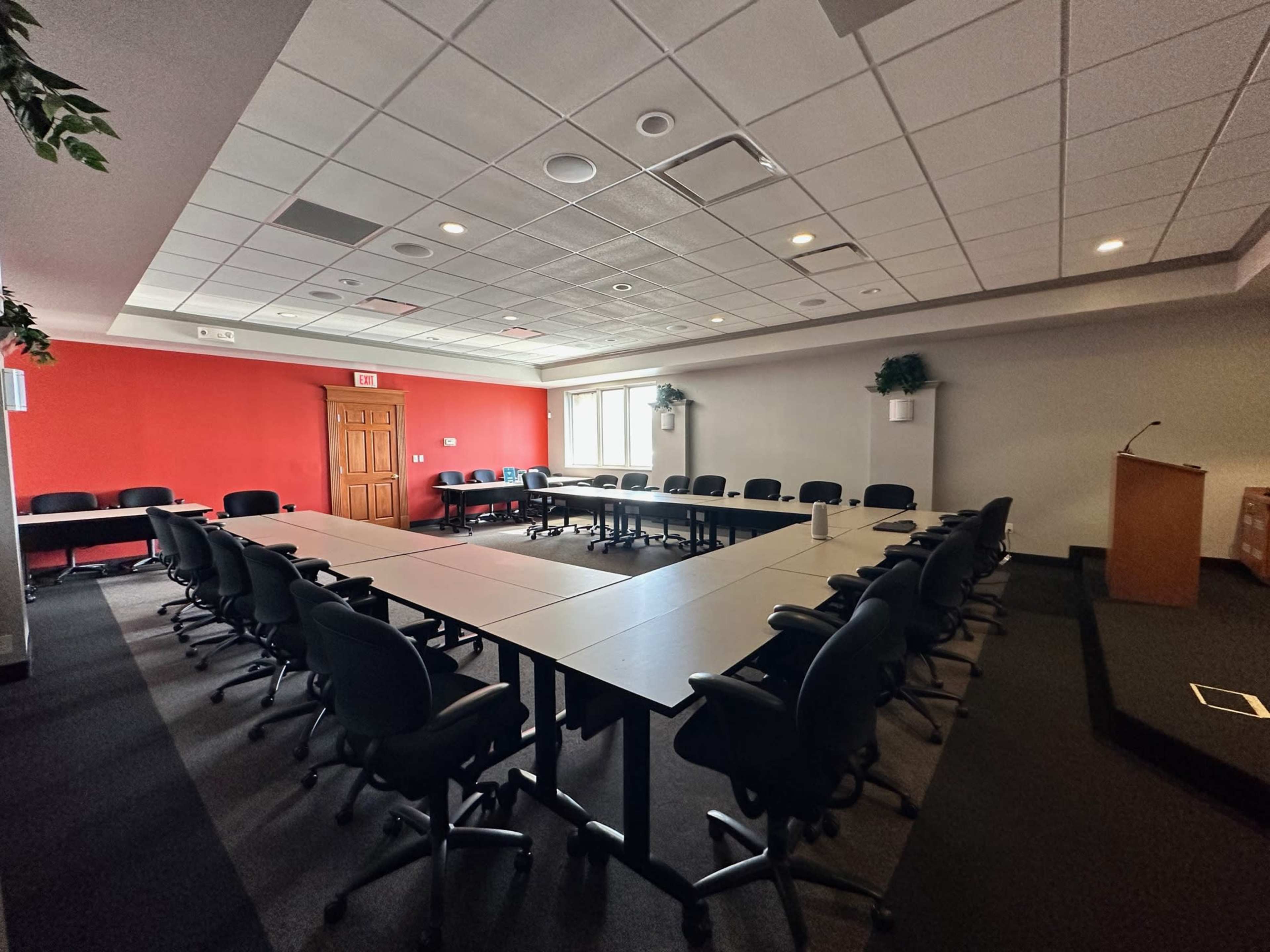 The image shows a large conference room arranged with a U-shaped seating layout around a central table, featuring a red accent wall and several windows.