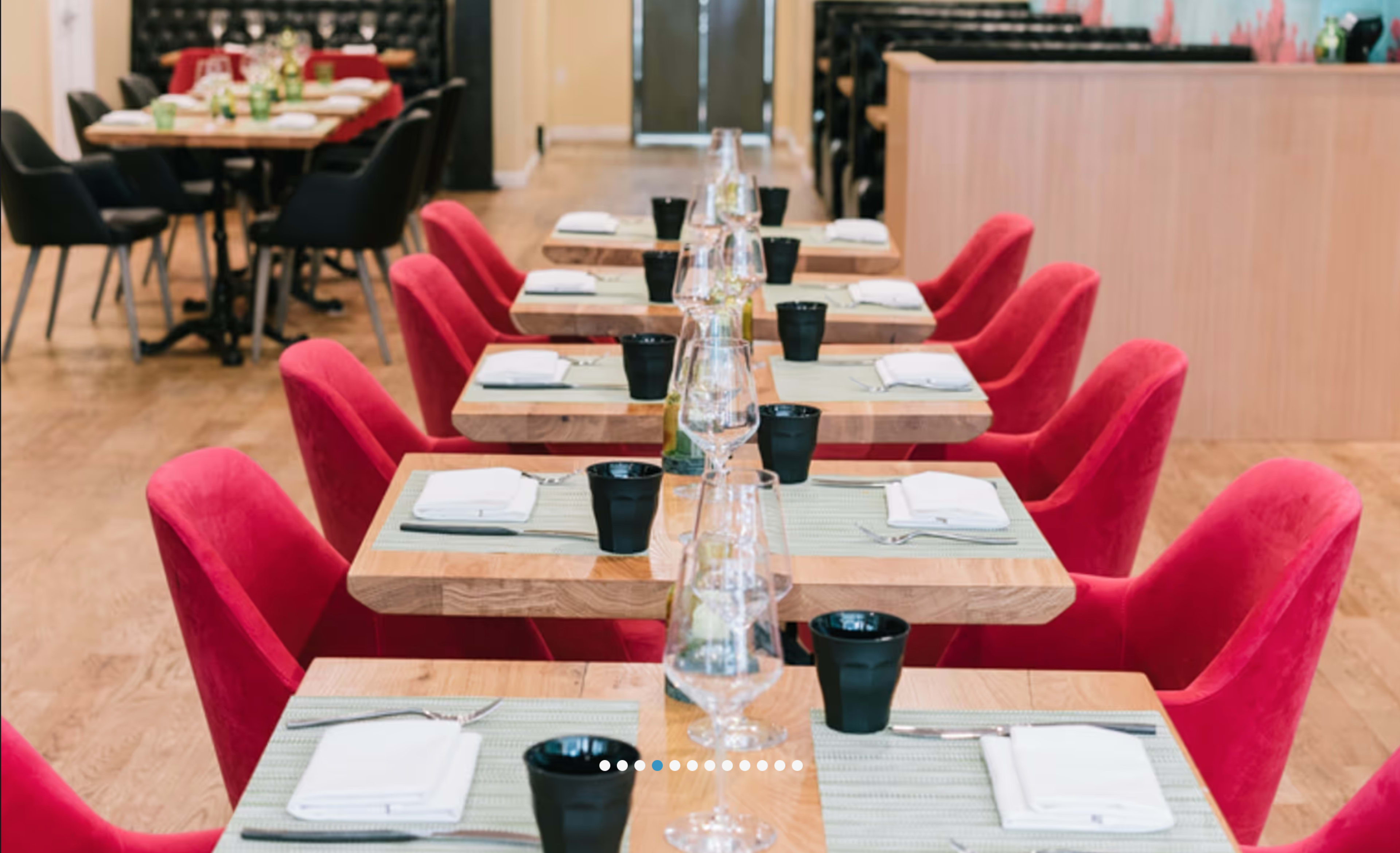 The image shows a dining area with rows of wooden tables set with white plates, glasses, and black cups, surrounded by red chairs.