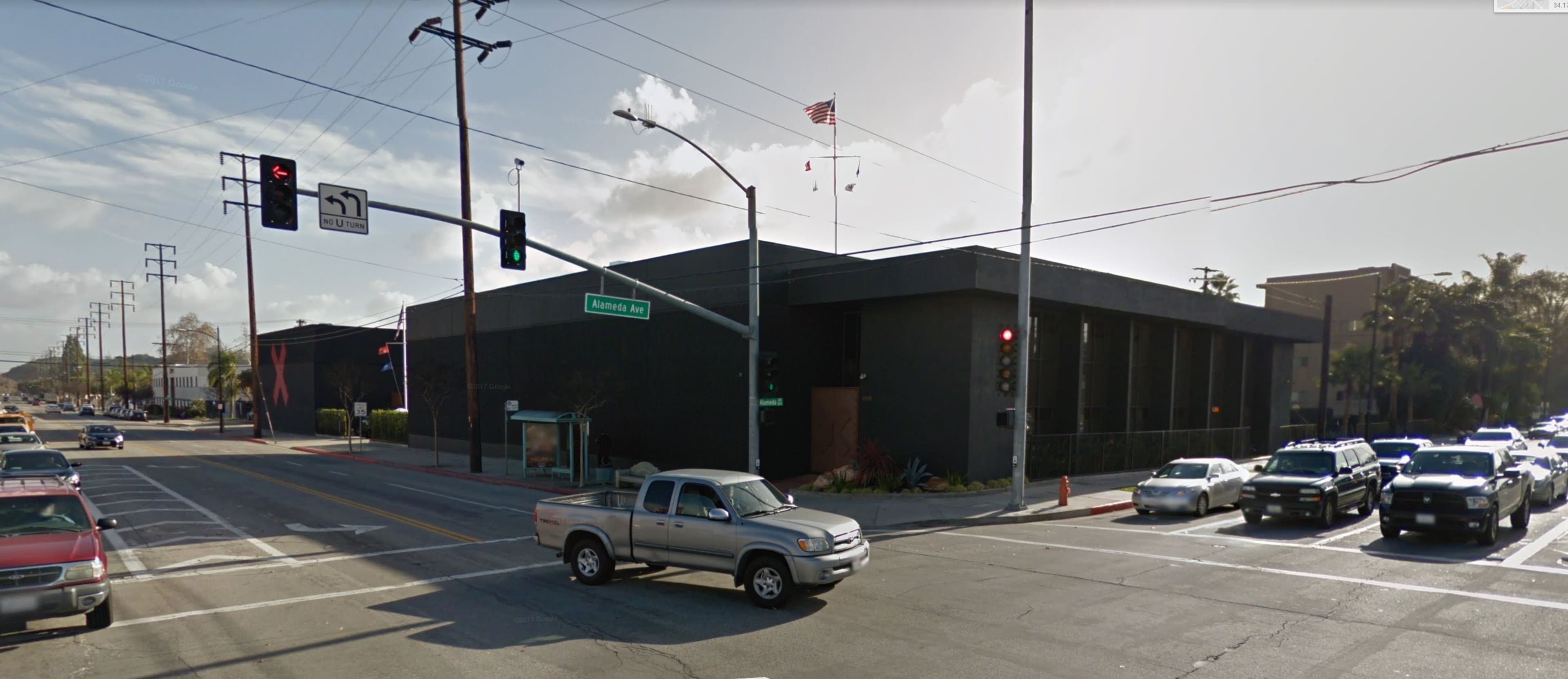 A street intersection with traffic lights and various vehicles, including a pickup truck, near a modern black building and a flagpole.