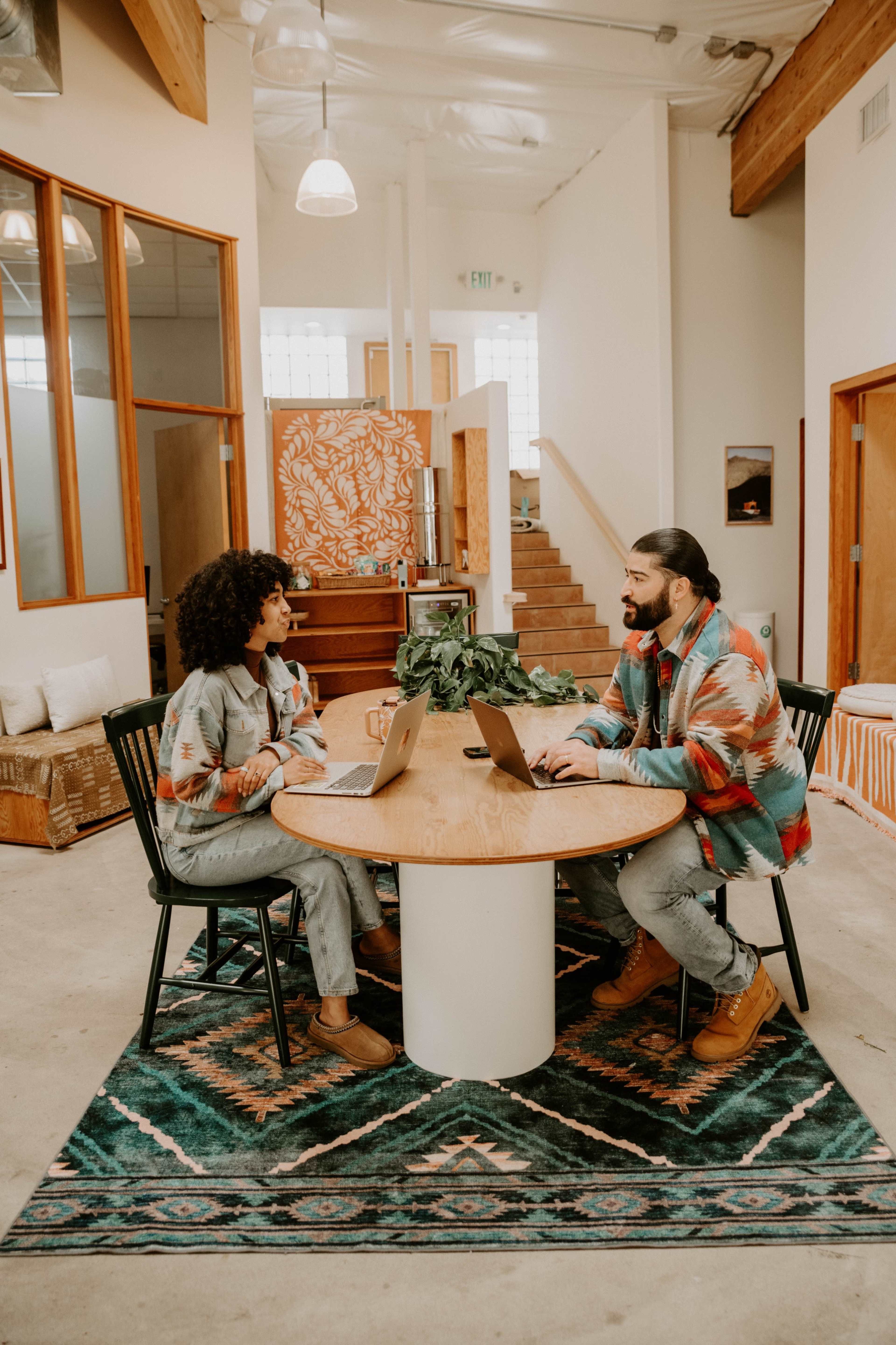 Two individuals are sitting at a round table in a modern workspace, each using a laptop, with a staircase and decorative wall art in the background.