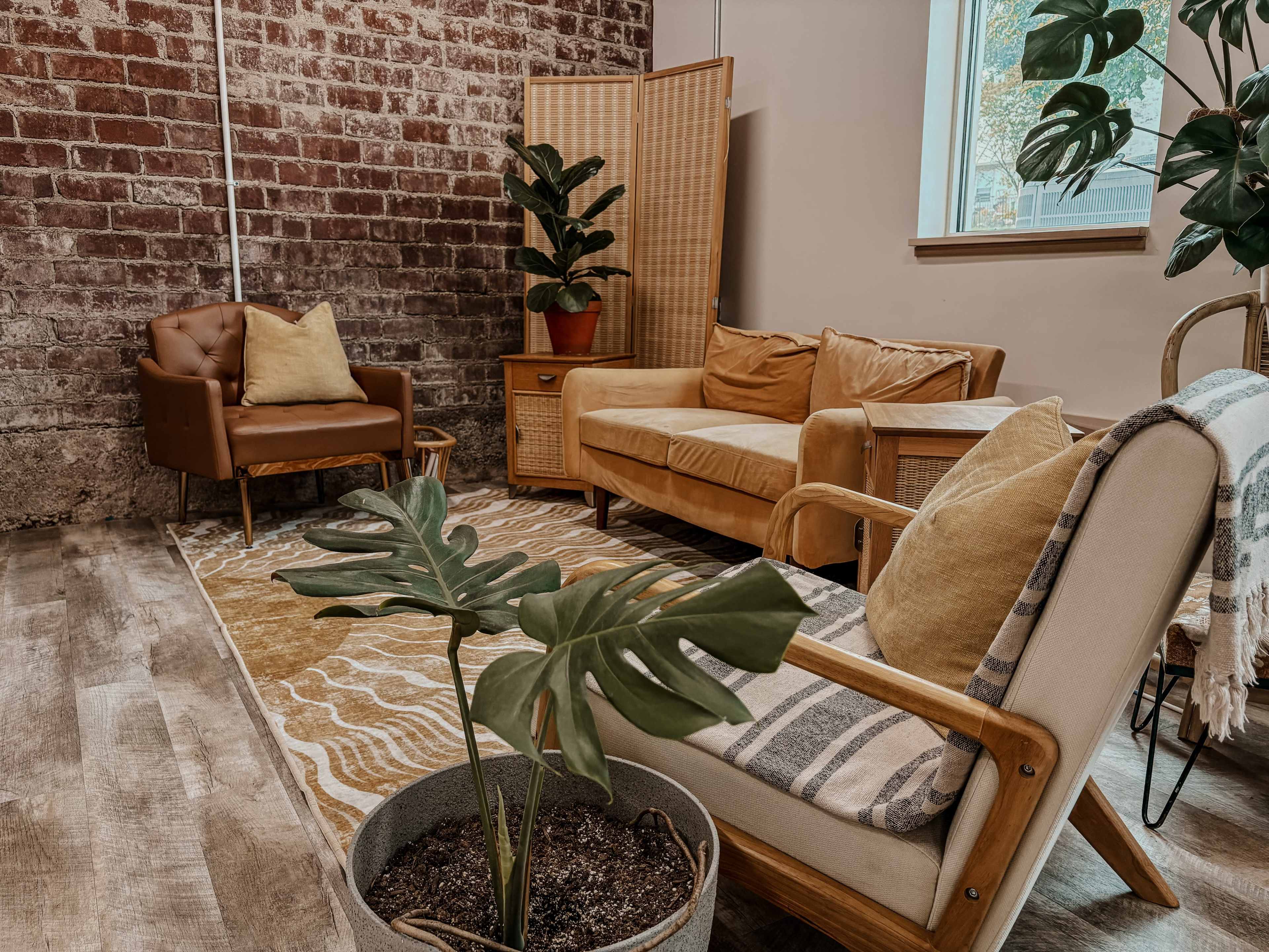 A cozy seating area features two armchairs and a sofa, surrounded by plants and a patterned rug against a brick wall.
