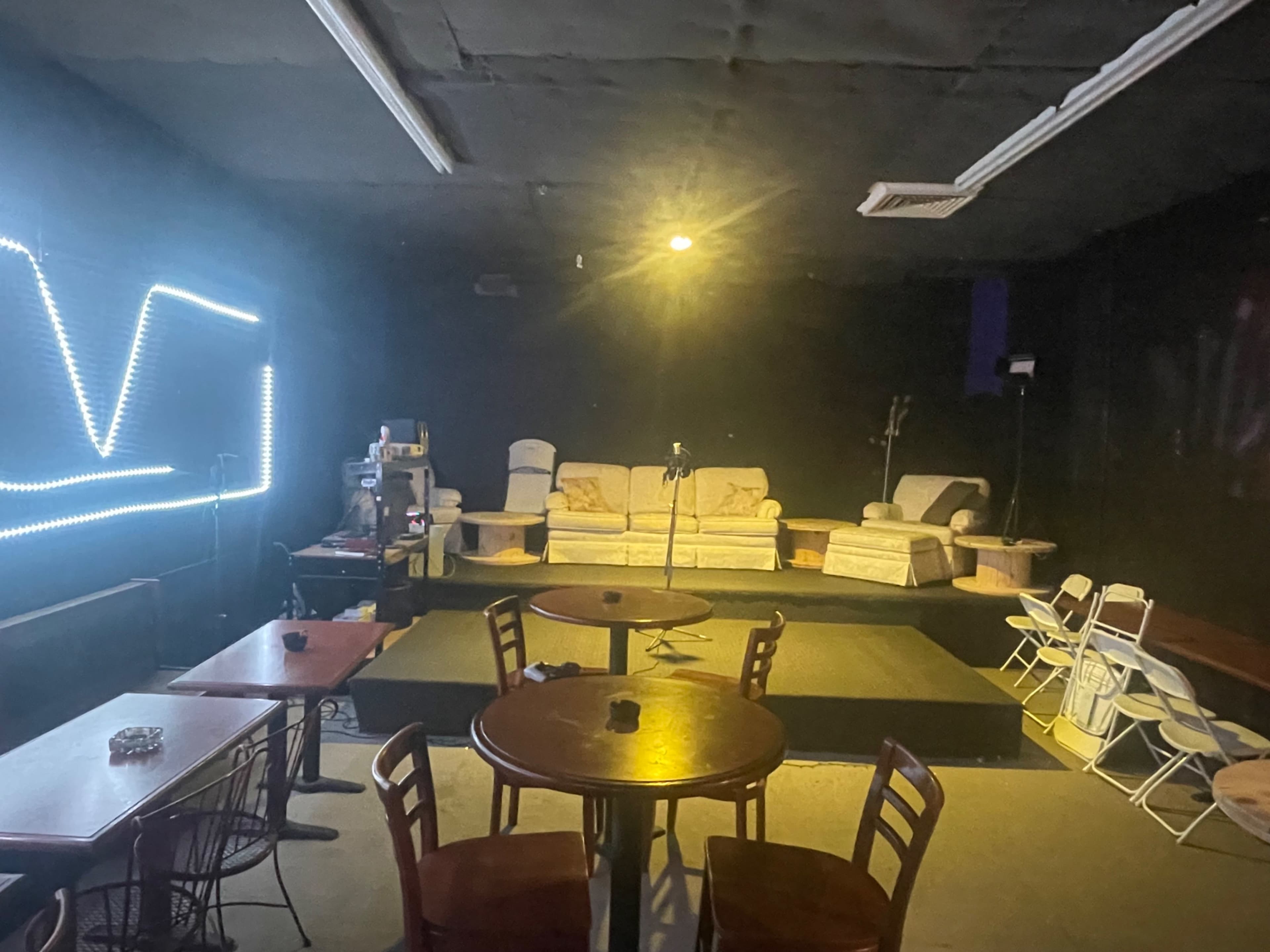 The image shows a dimly lit performance space with two couches on a small stage area, surrounded by tables and chairs.