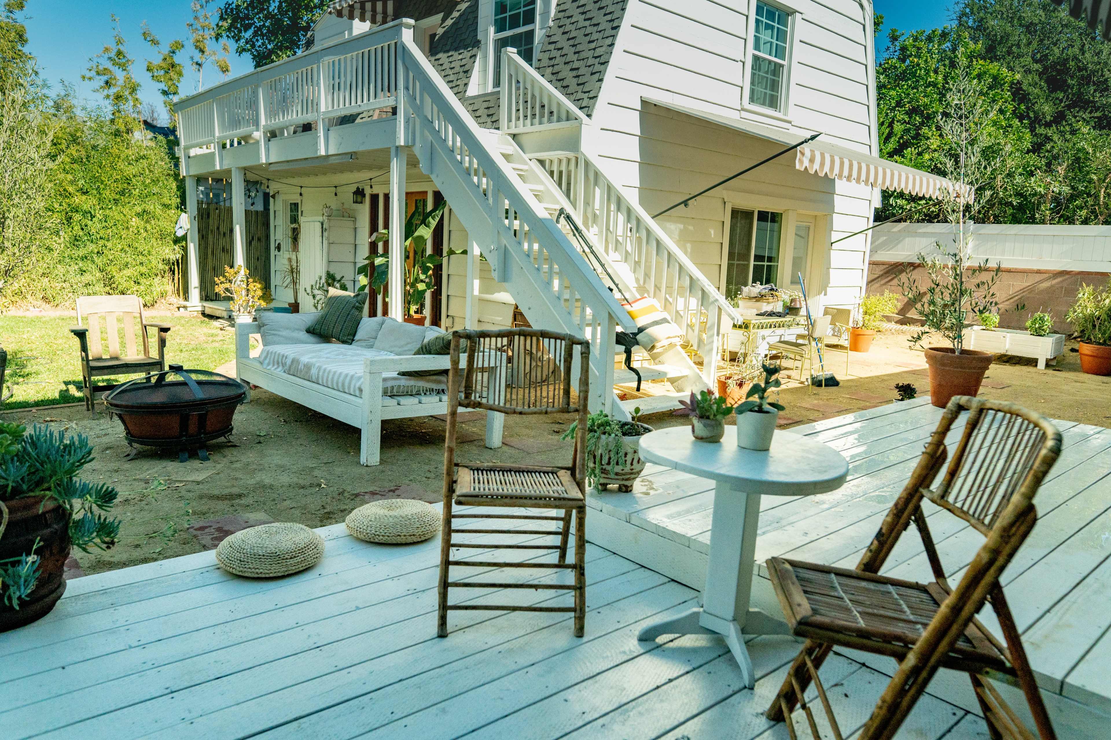 A bright outdoor area features a white wooden deck with a small table and two chairs, leading to a house with a staircase and a cozy seating area underneath.