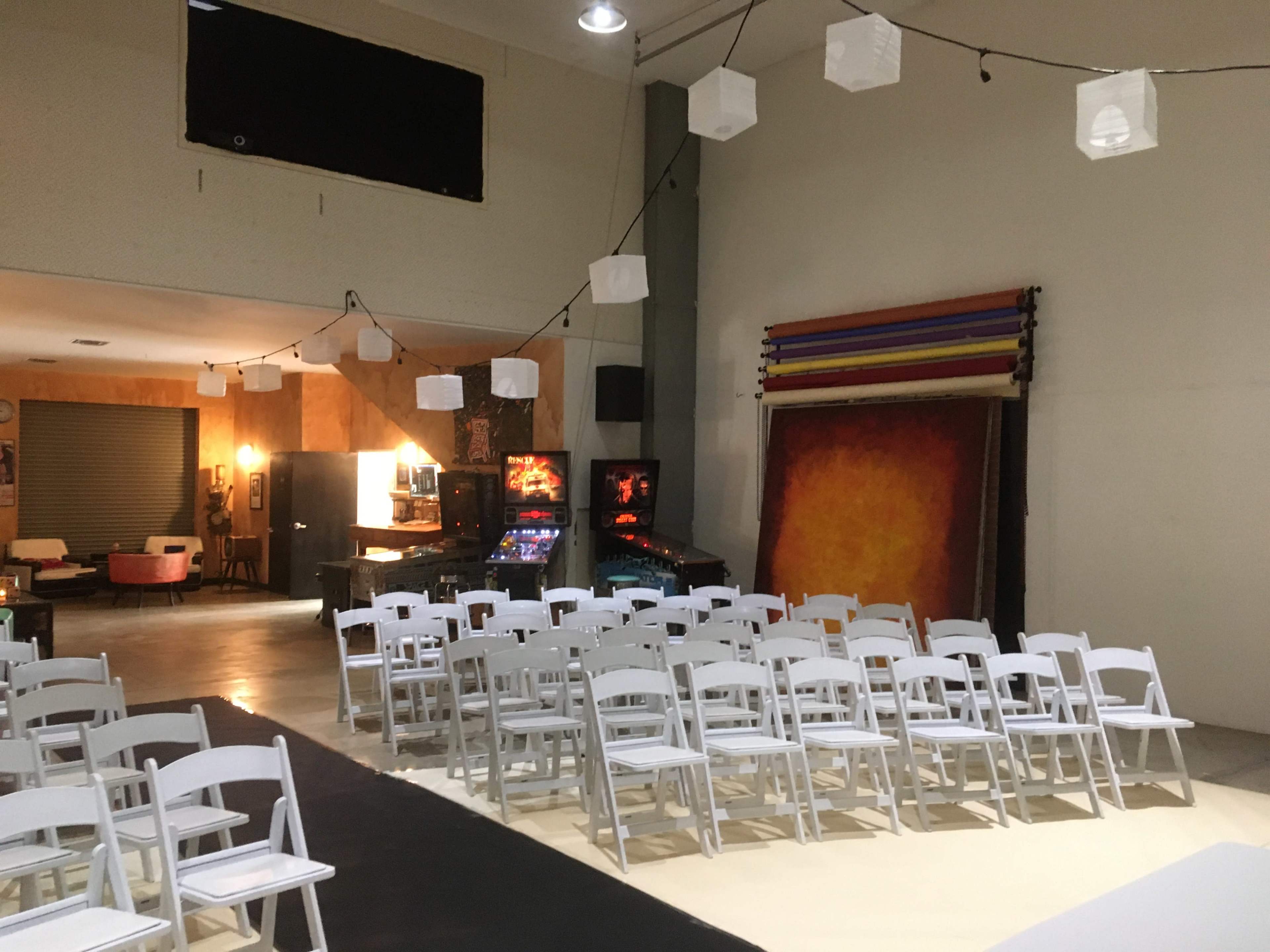 A spacious room set up with white folding chairs arranged for an event, with pinball machines and artwork displayed in the background.