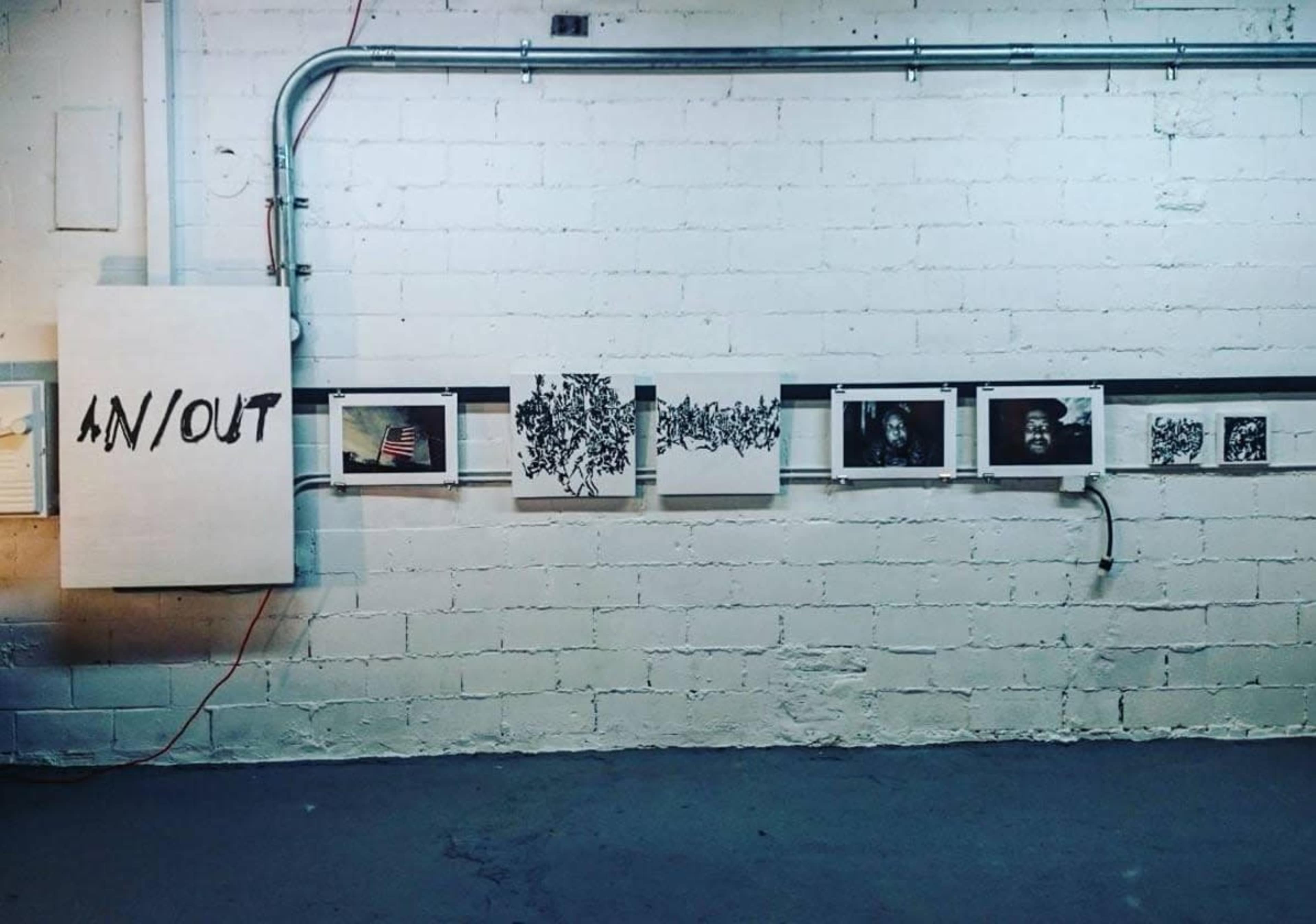 The image shows a gallery wall with a large canvas labeled "IN/OUT" and several framed artworks displayed beneath it.