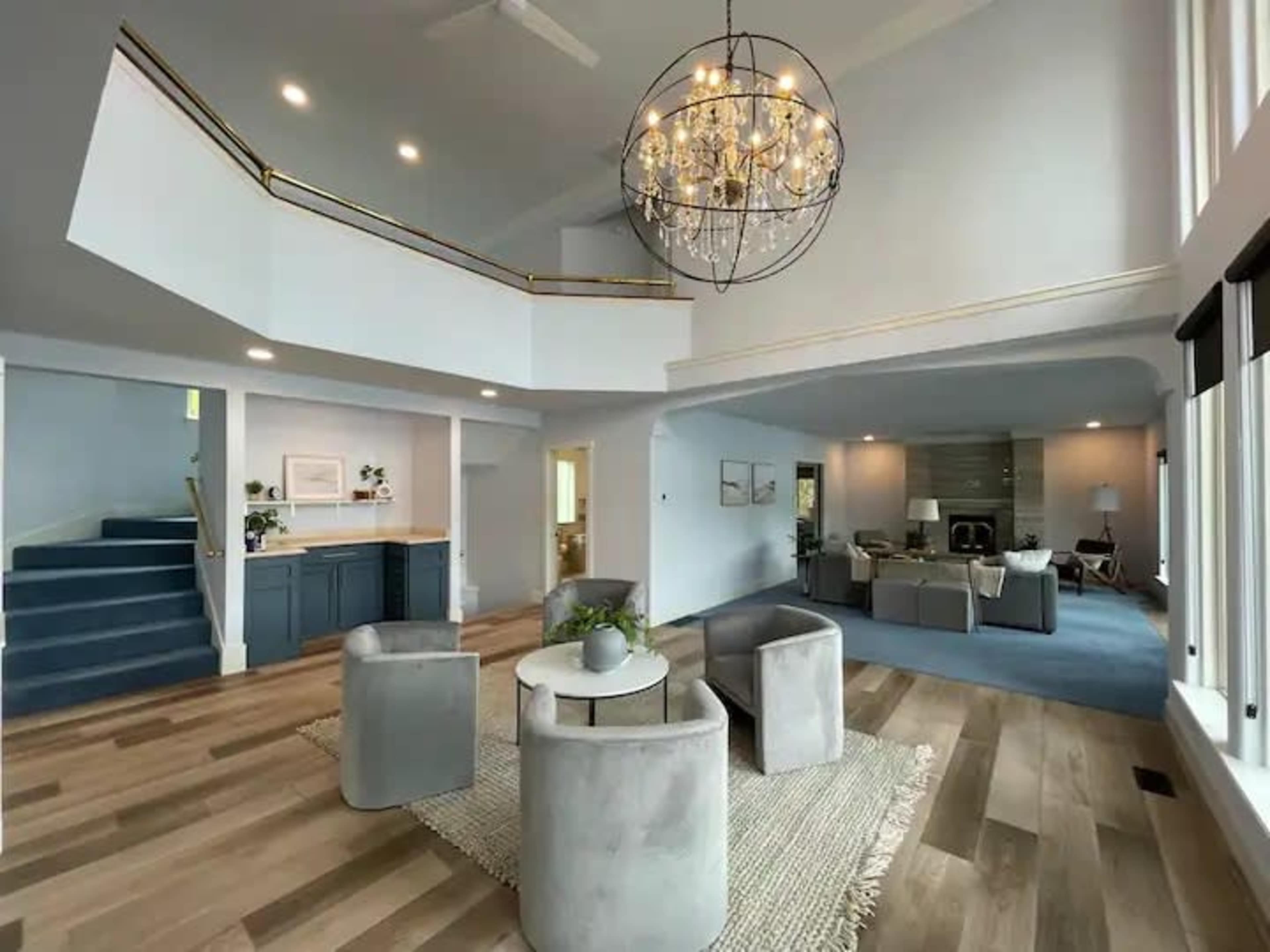 A spacious living area with a modern chandelier, a round table surrounded by four chairs, and a staircase leading to an upper level, all featuring light-colored walls and wooden flooring.