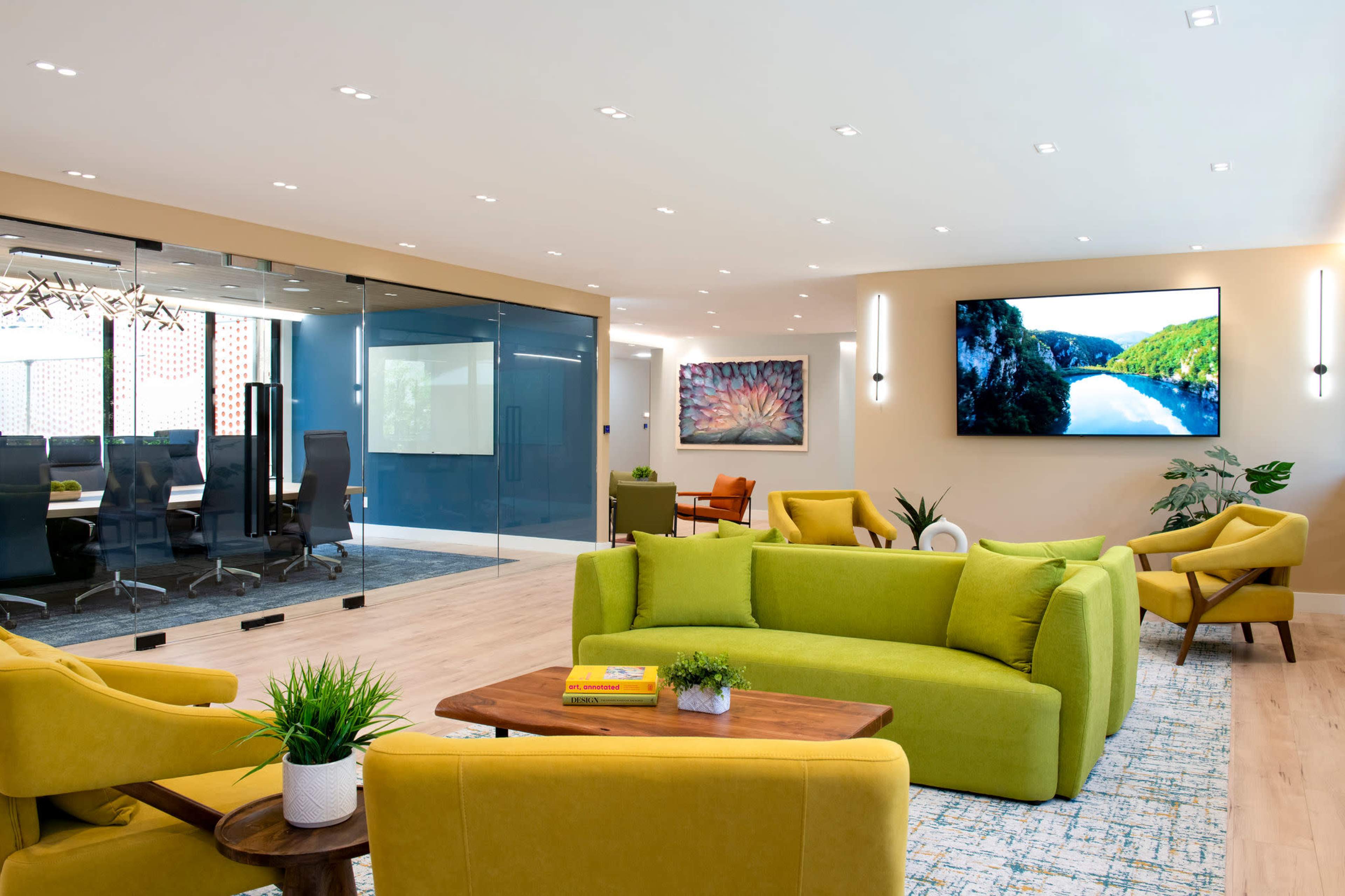The image shows a modern office interior featuring green sofas, a wooden coffee table, and a large screen displaying a landscape, with a glass-walled conference room in the background.