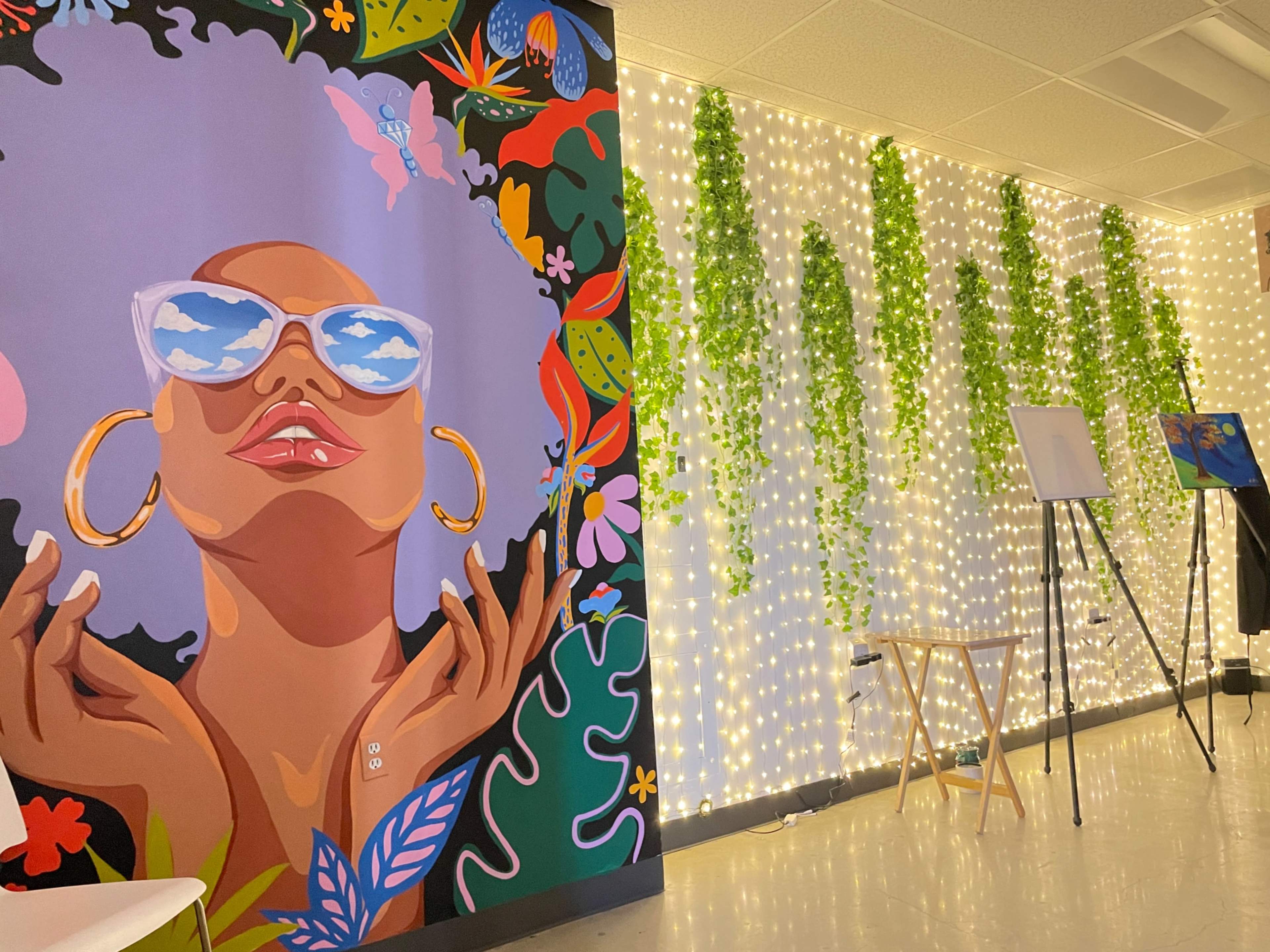 A vibrant mural of a woman with sunglasses on one wall, while the opposite wall is adorned with string lights and hanging greenery.