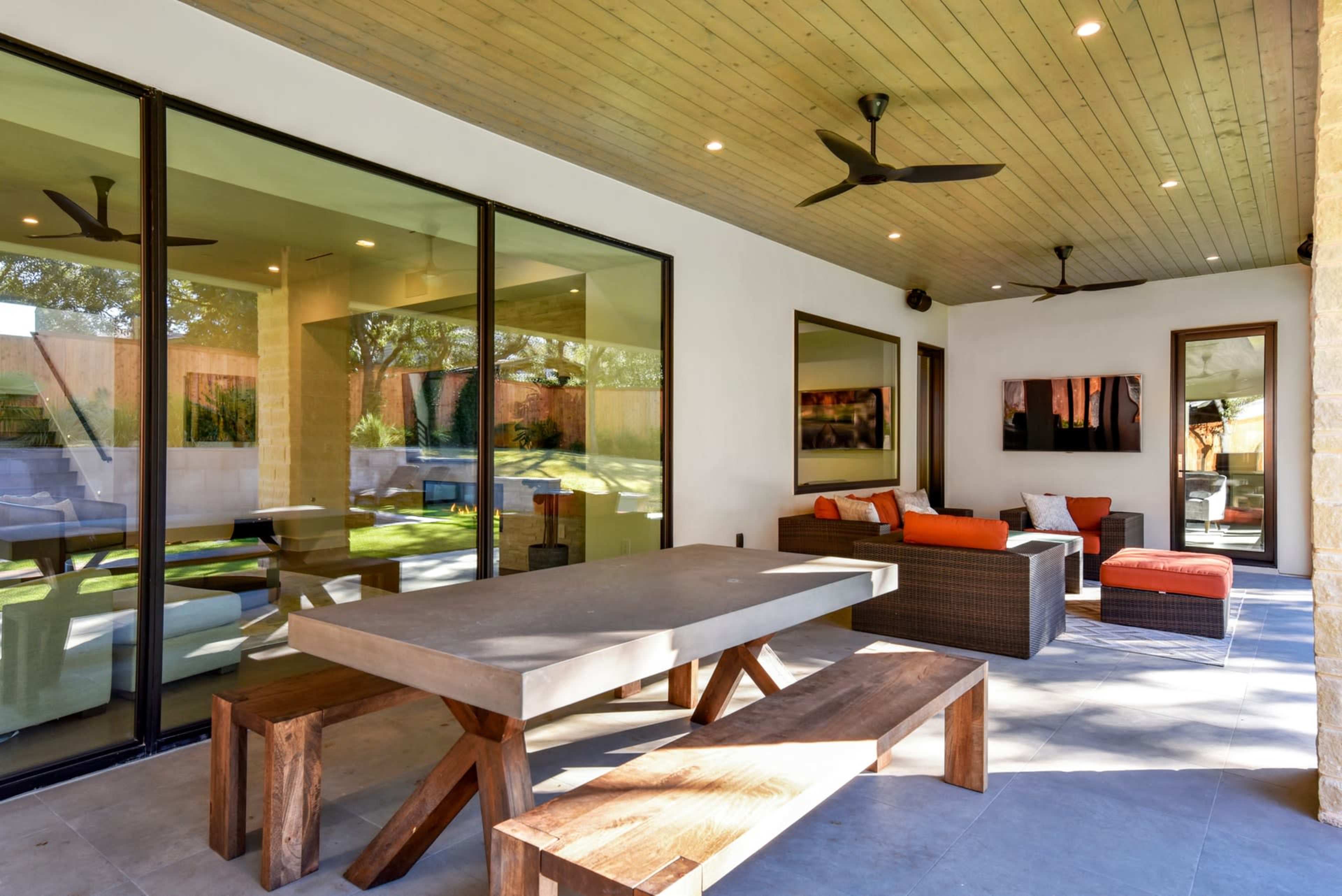 The image shows a spacious outdoor patio with a dining table, seating area, and large glass doors that open to a backyard with a pool.