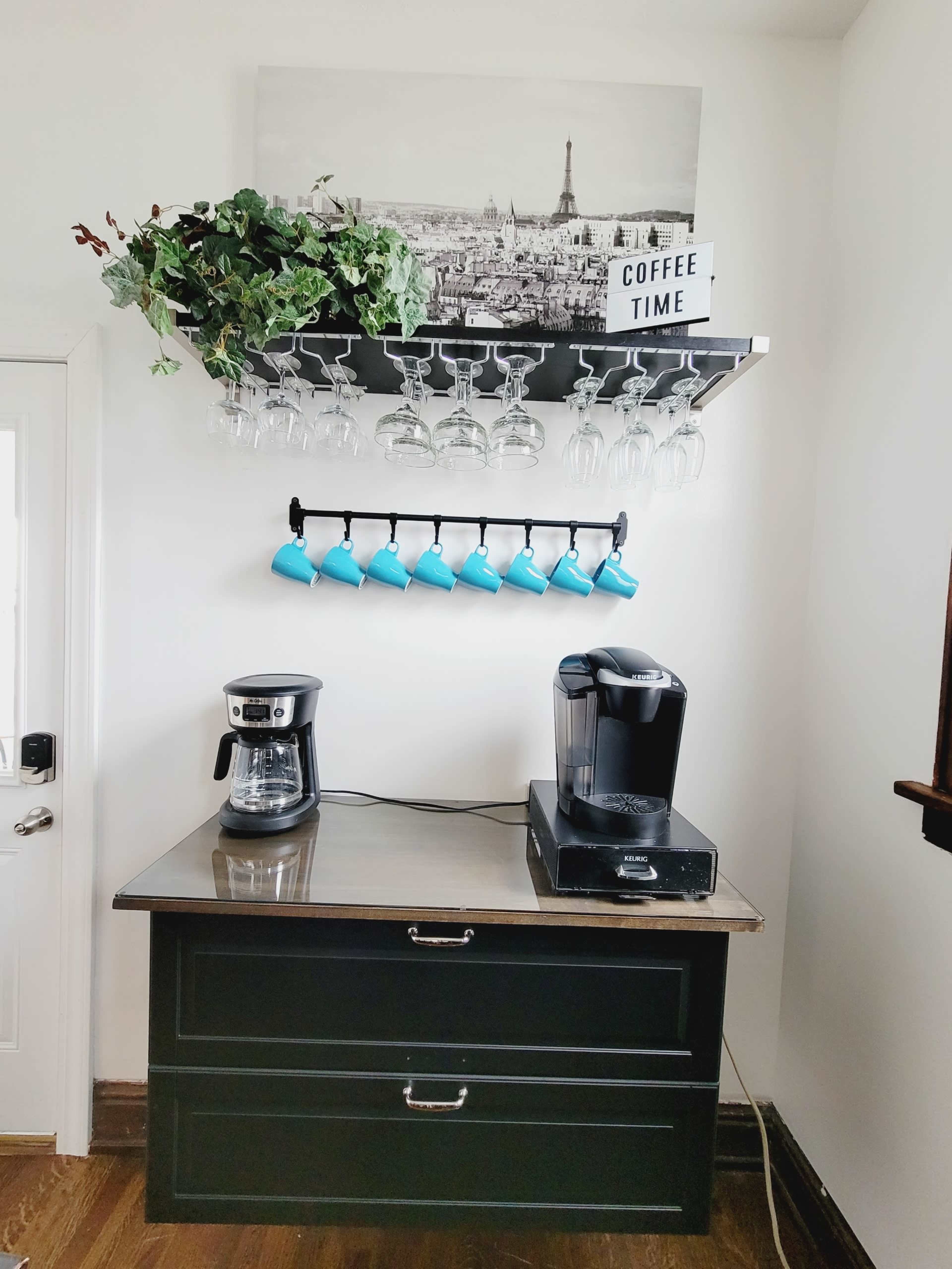A coffee station with a black countertop, a coffee maker, a Keurig machine, a shelf holding glasses, and blue mugs, along with a decorative plant and a sign that reads "COFFEE TIME."
