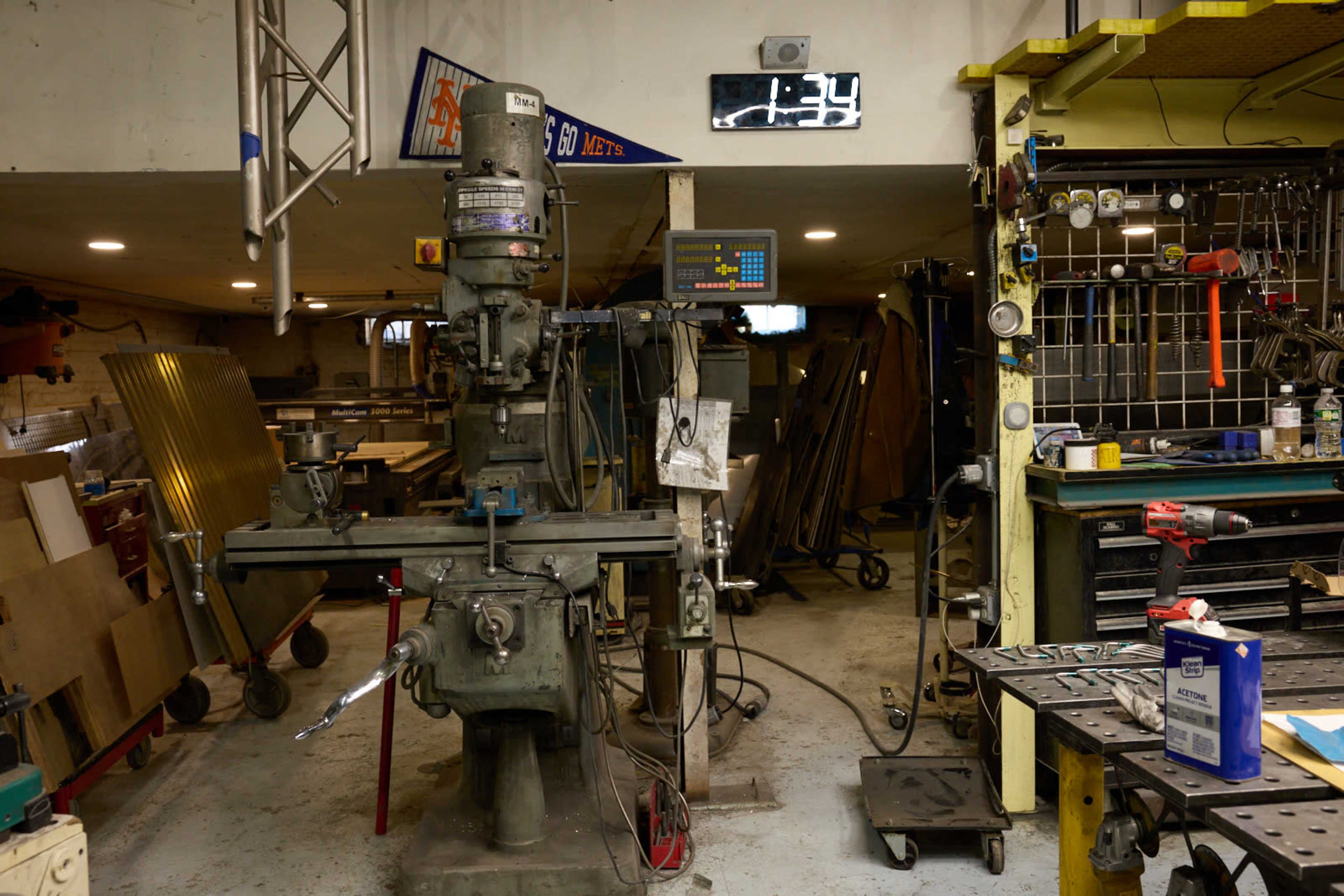 A milling machine is prominently positioned in a workshop filled with tools, equipment, and materials organized on shelves and tables.