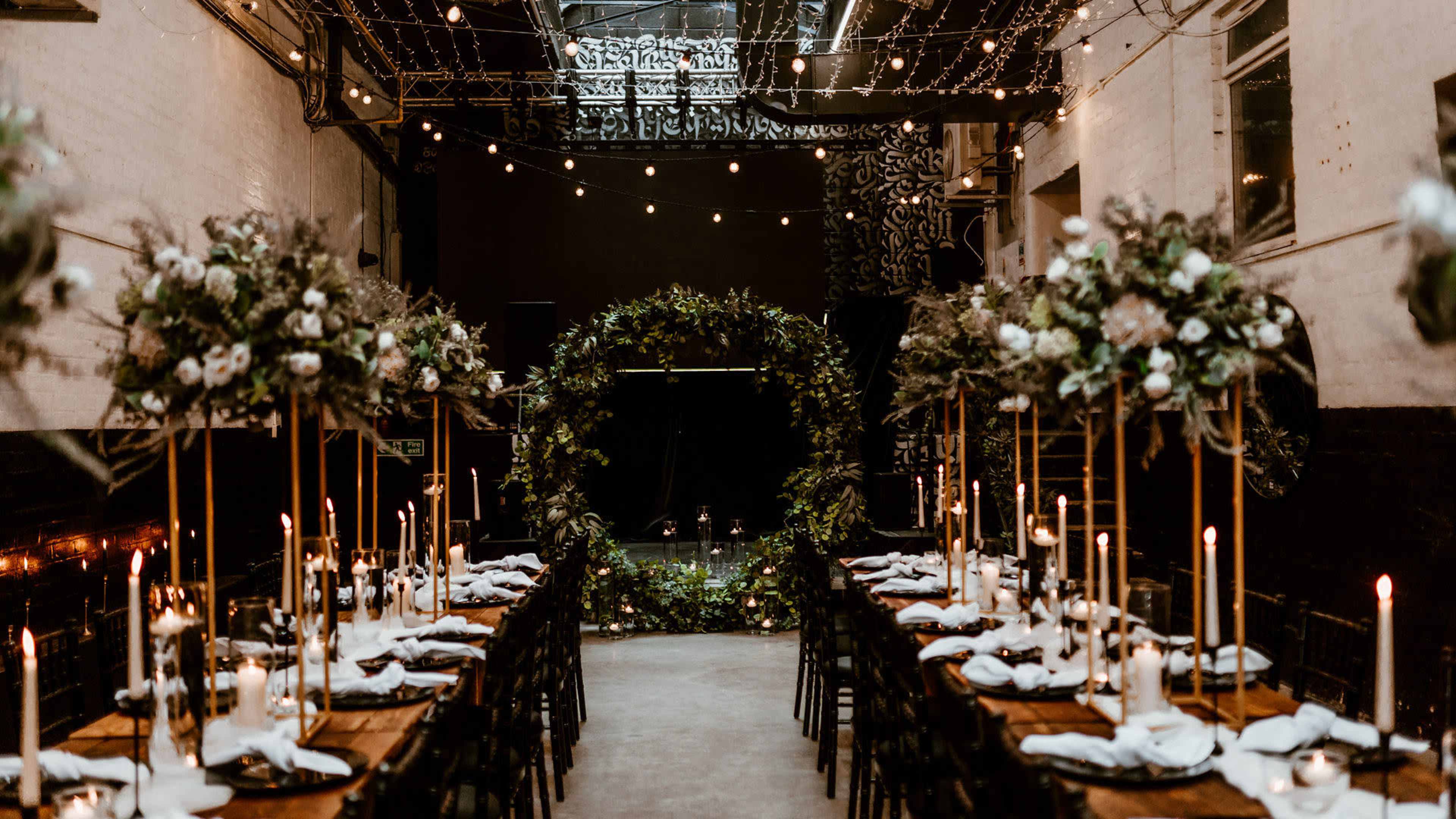 A decorated event space for a wedding, featuring long wooden tables set with white tableware, tall floral arrangements, and an archway adorned with greenery at one end.