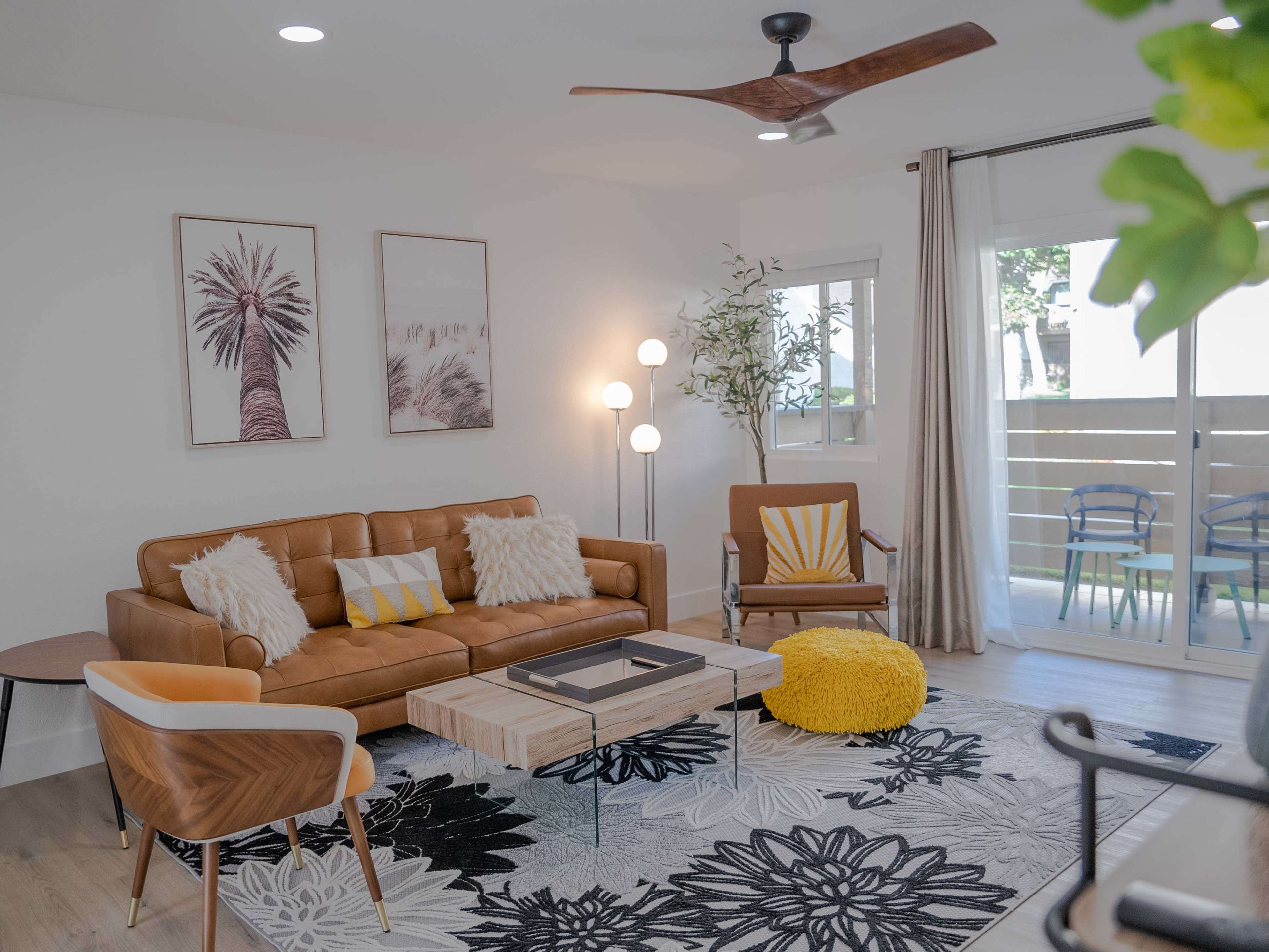 The living room features a brown leather sofa, a wooden coffee table, a patterned rug, and large windows with sliding glass doors.