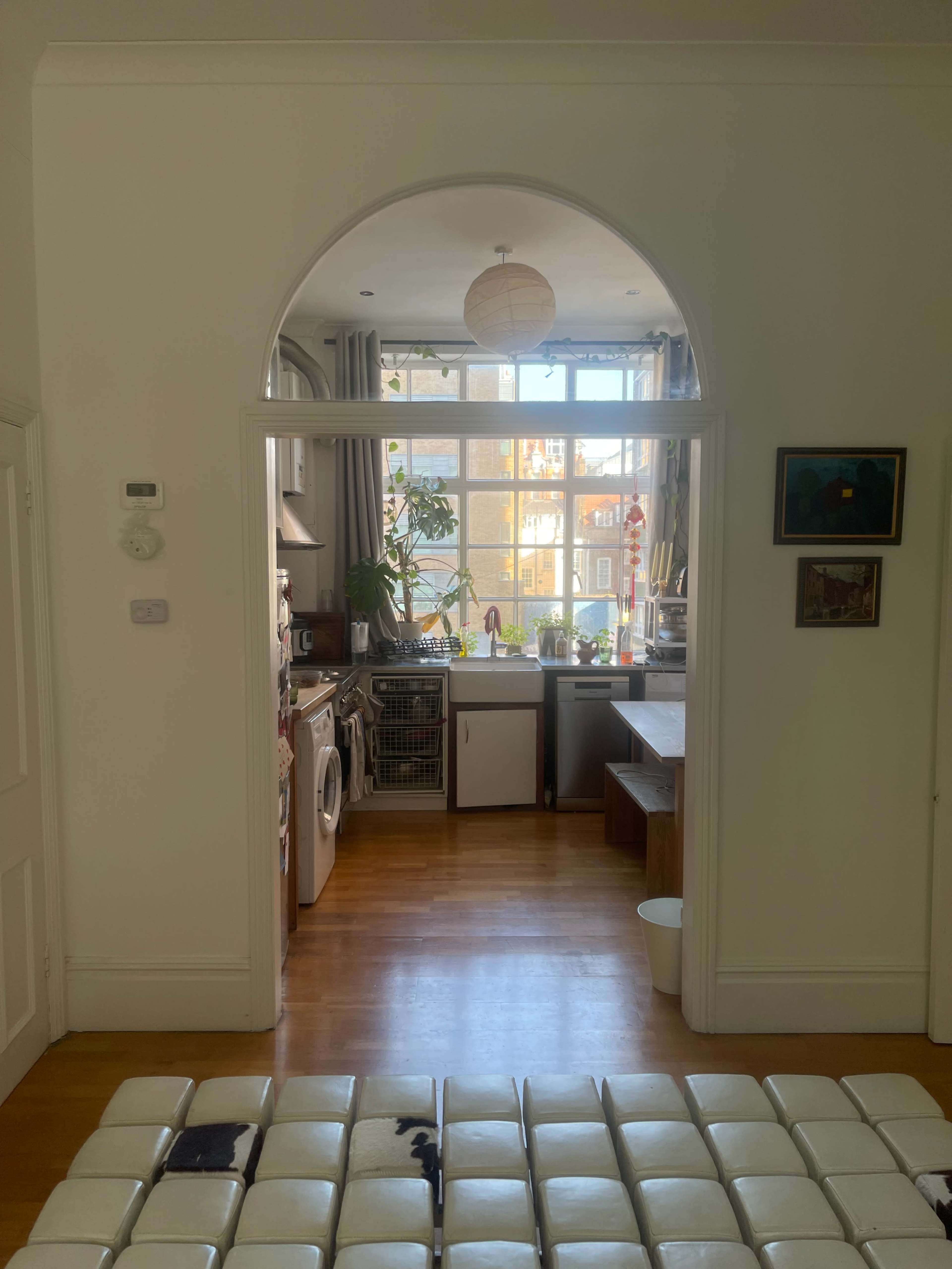 A kitchen is visible through an arched doorway, showcasing appliances, cabinets, and a dining area with large windows allowing natural light.