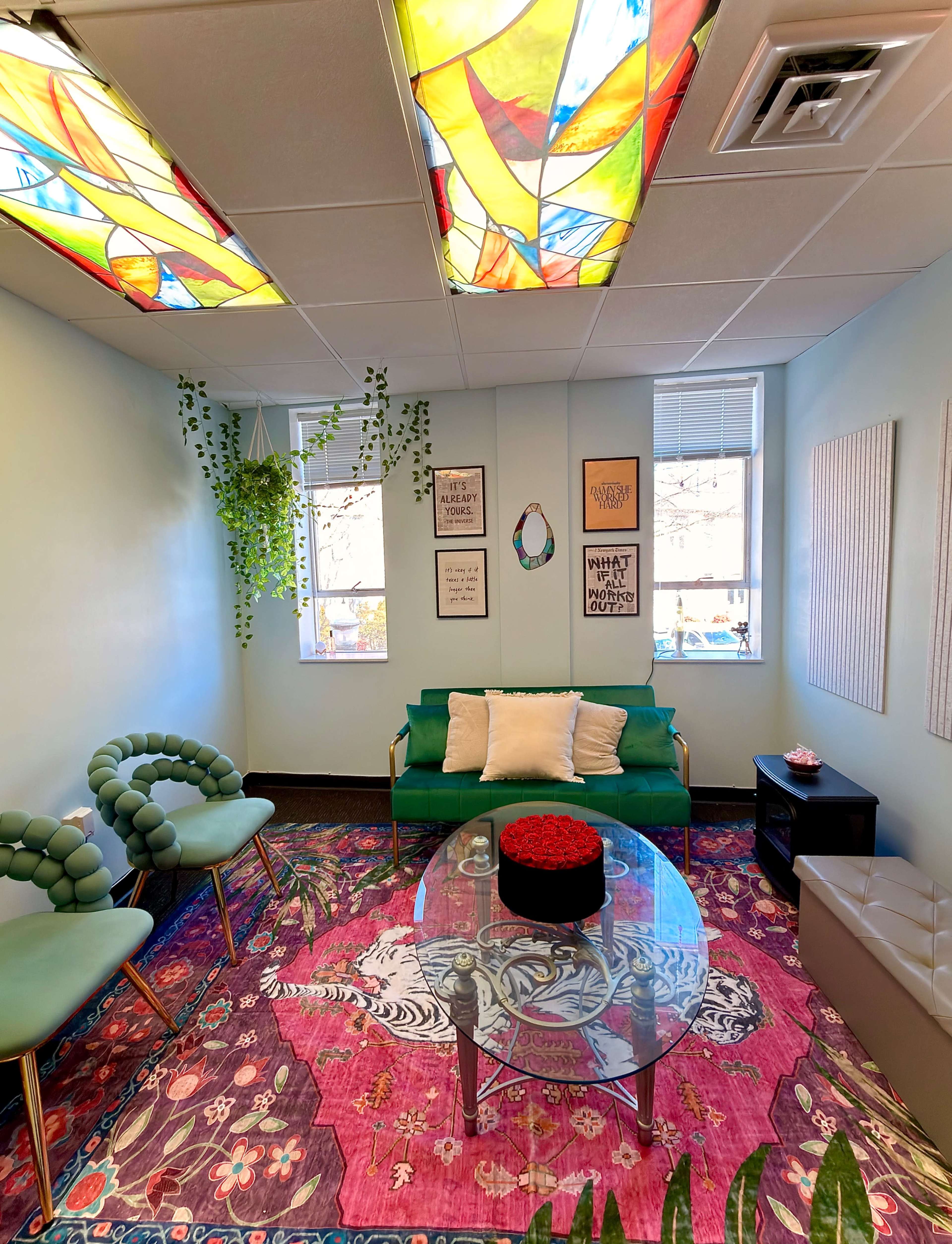 The room features a green couch, two green chairs, a round glass table on a vibrant patterned rug, and stained glass windows above, creating a bright and inviting atmosphere.