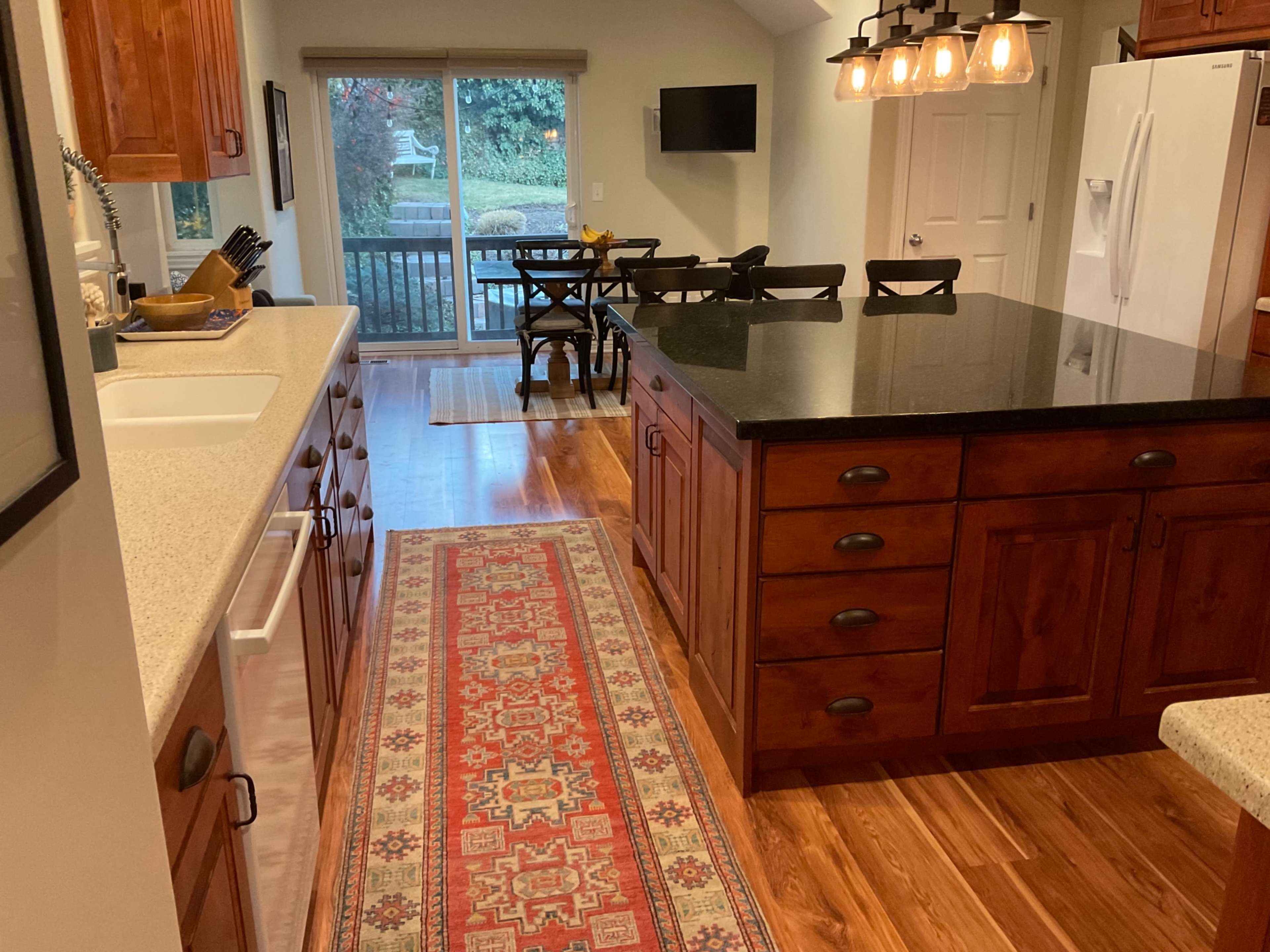 A modern kitchen features wooden cabinets, a large island with a black countertop, and a dining area visible through the sliding glass door.