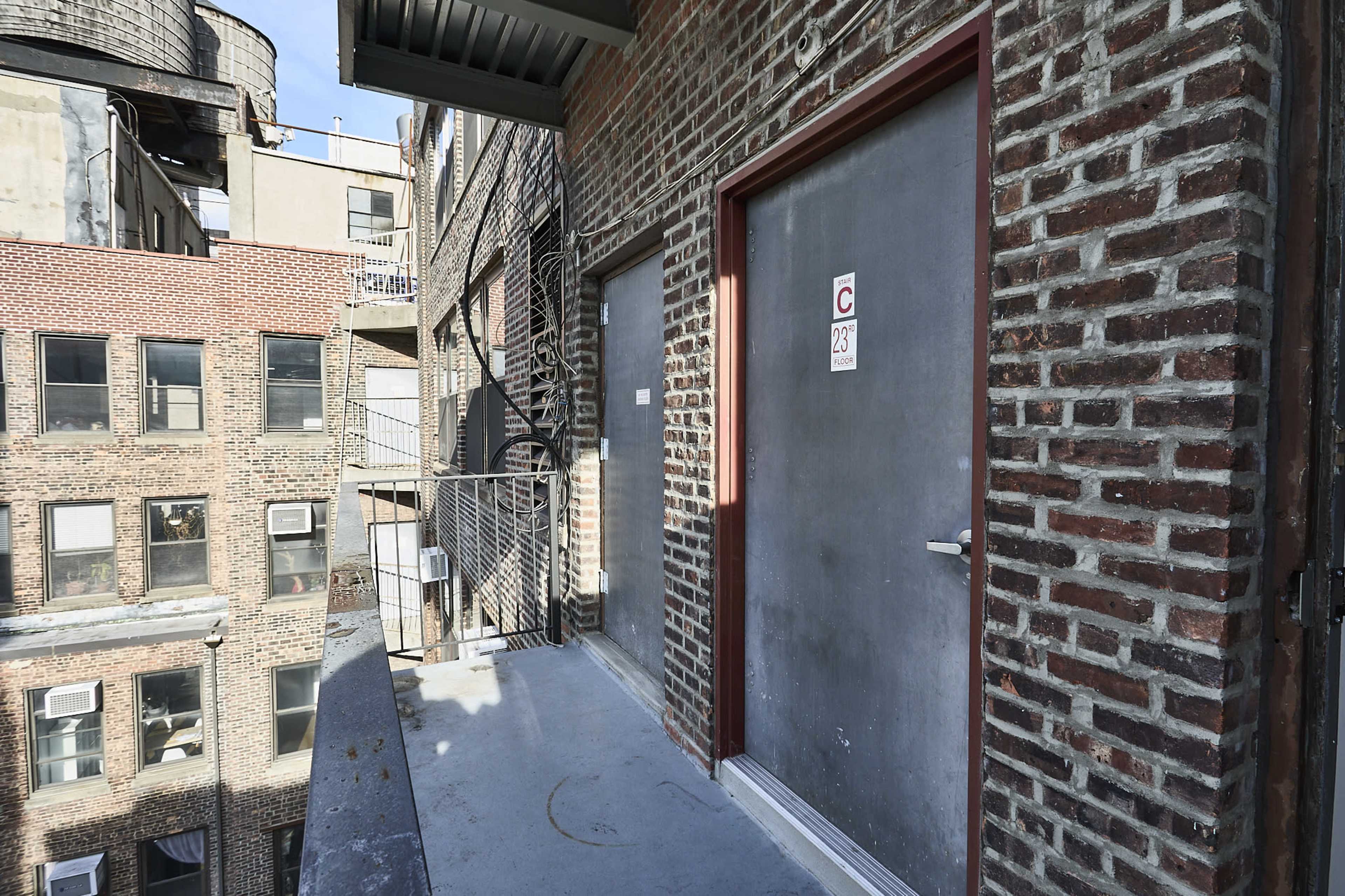 The image shows a narrow balcony overlooking a brick-walled courtyard, featuring two closed doors and metal railings.