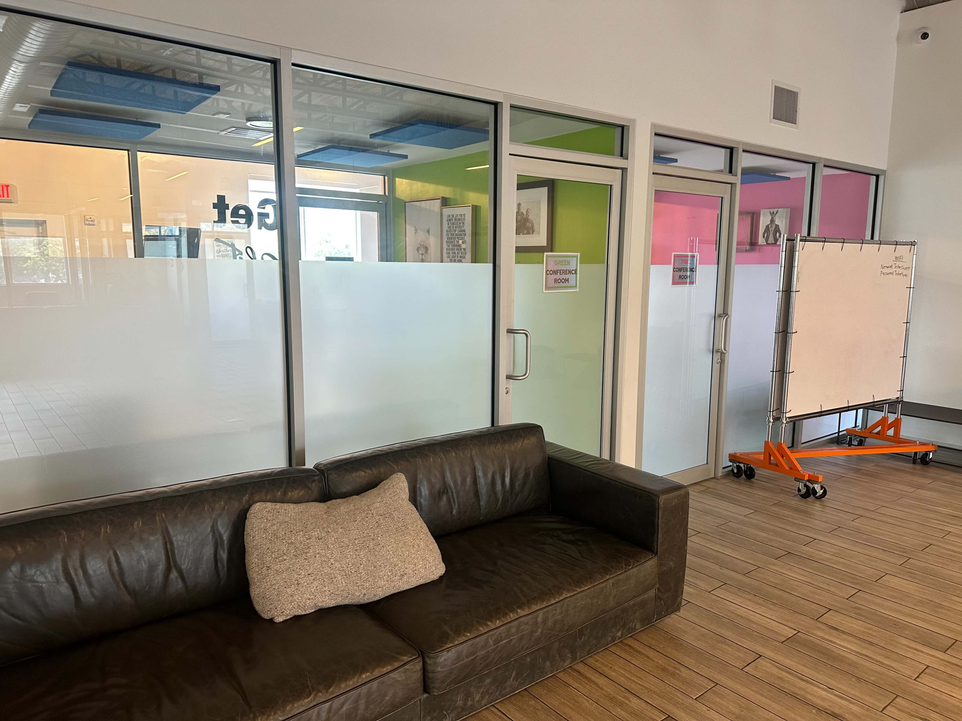 A brown leather couch with a decorative pillow is positioned near a glass wall with colorful panels and a rolling whiteboard in a well-lit indoor space.