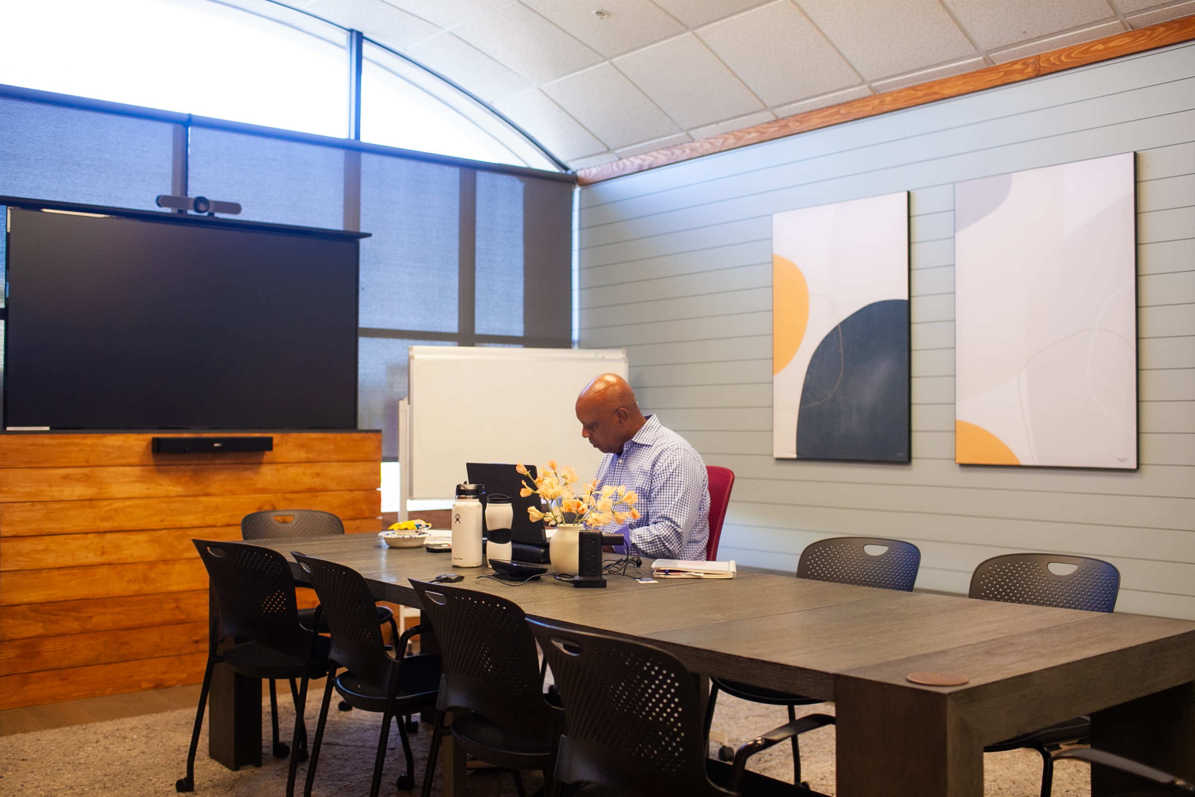 A man works at a table in a meeting room featuring a large screen, whiteboard, and abstract wall art.