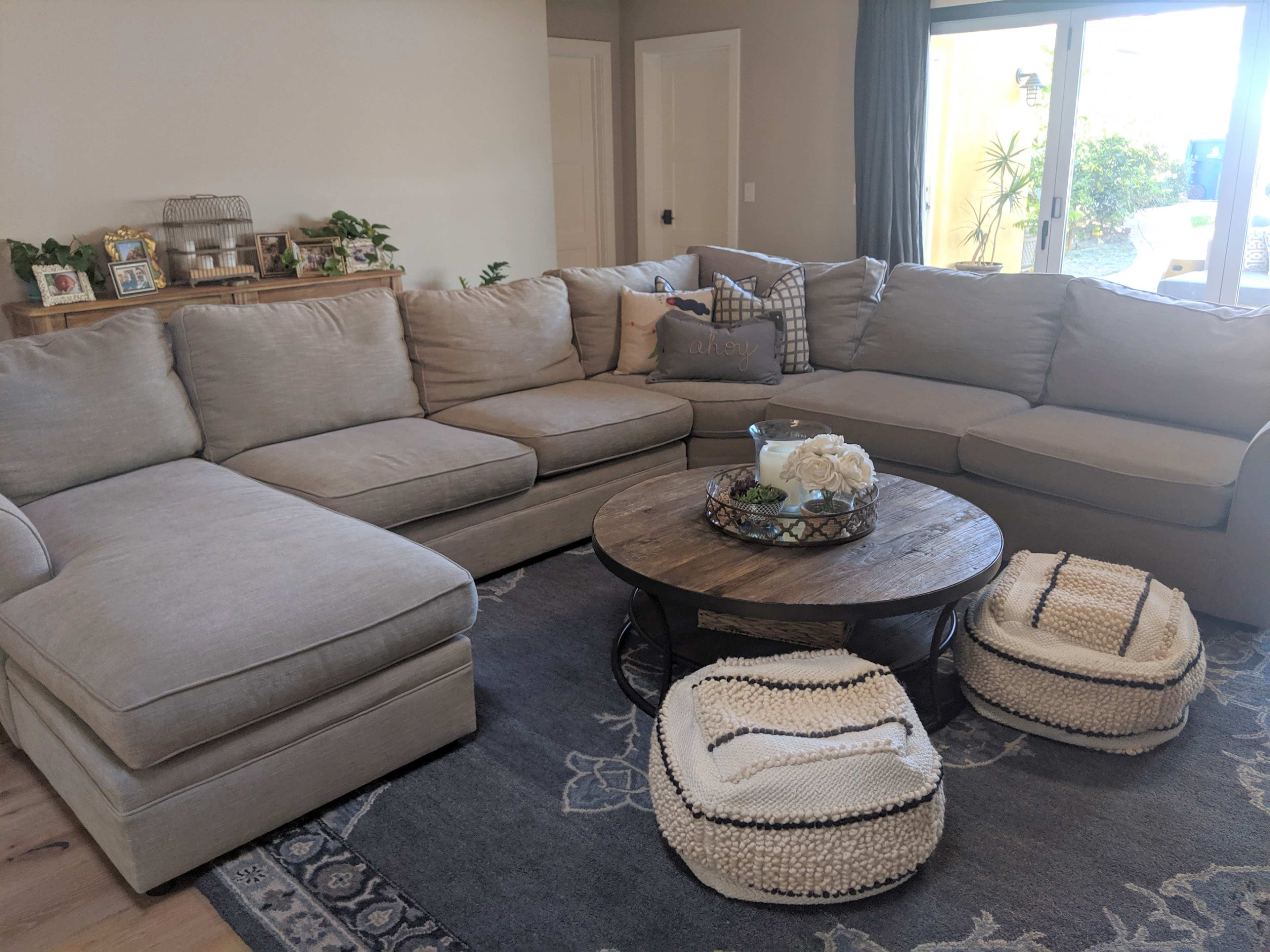 The image shows a spacious living room featuring a large sectional sofa, a round coffee table, and two woven poufs on a decorative rug.