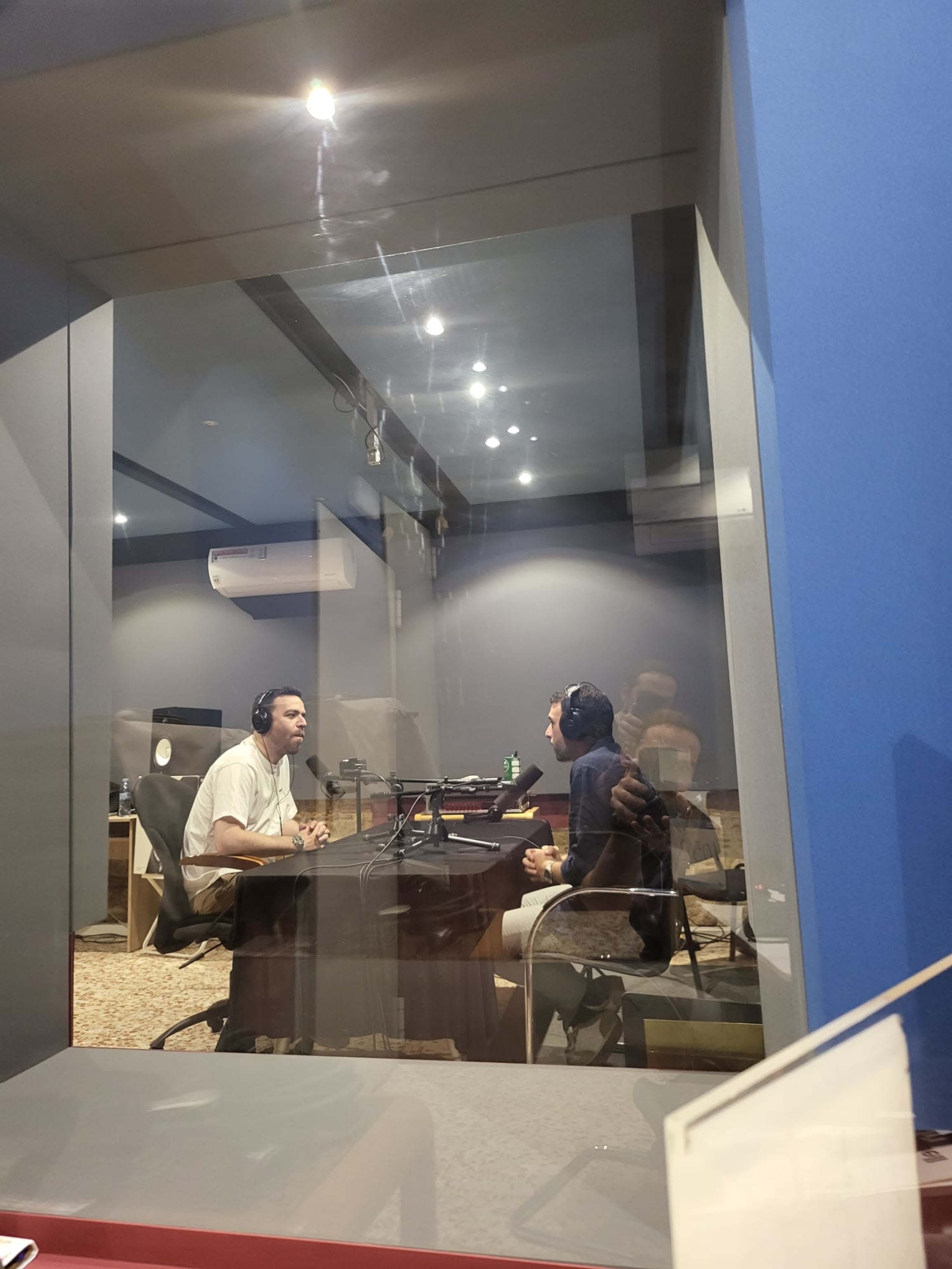 Two individuals are seated at a table in a recording studio, wearing headphones and speaking to each other.