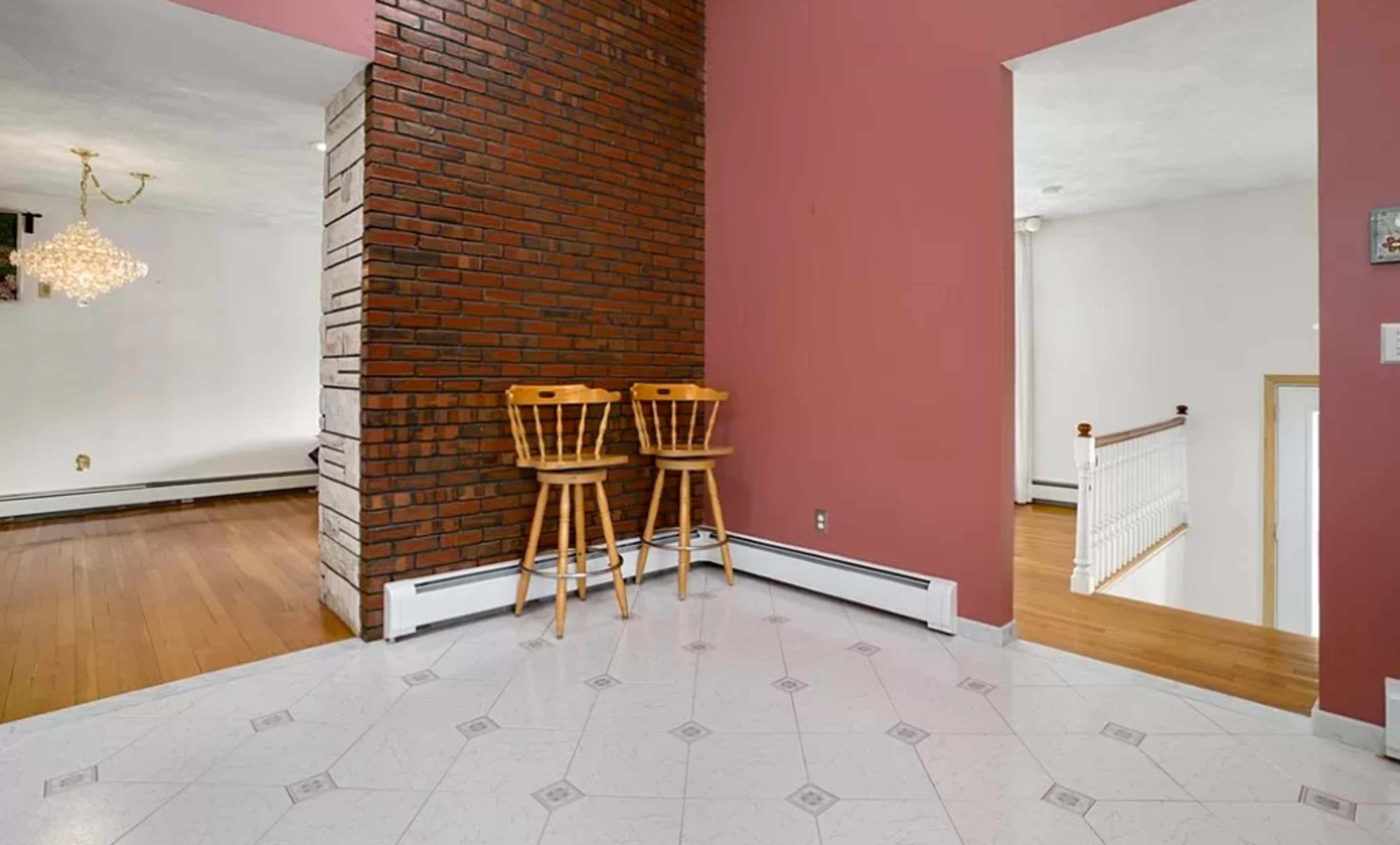 The image shows a room with a brick wall, two wooden stools, and an entrance leading to another area with hardwood flooring.
