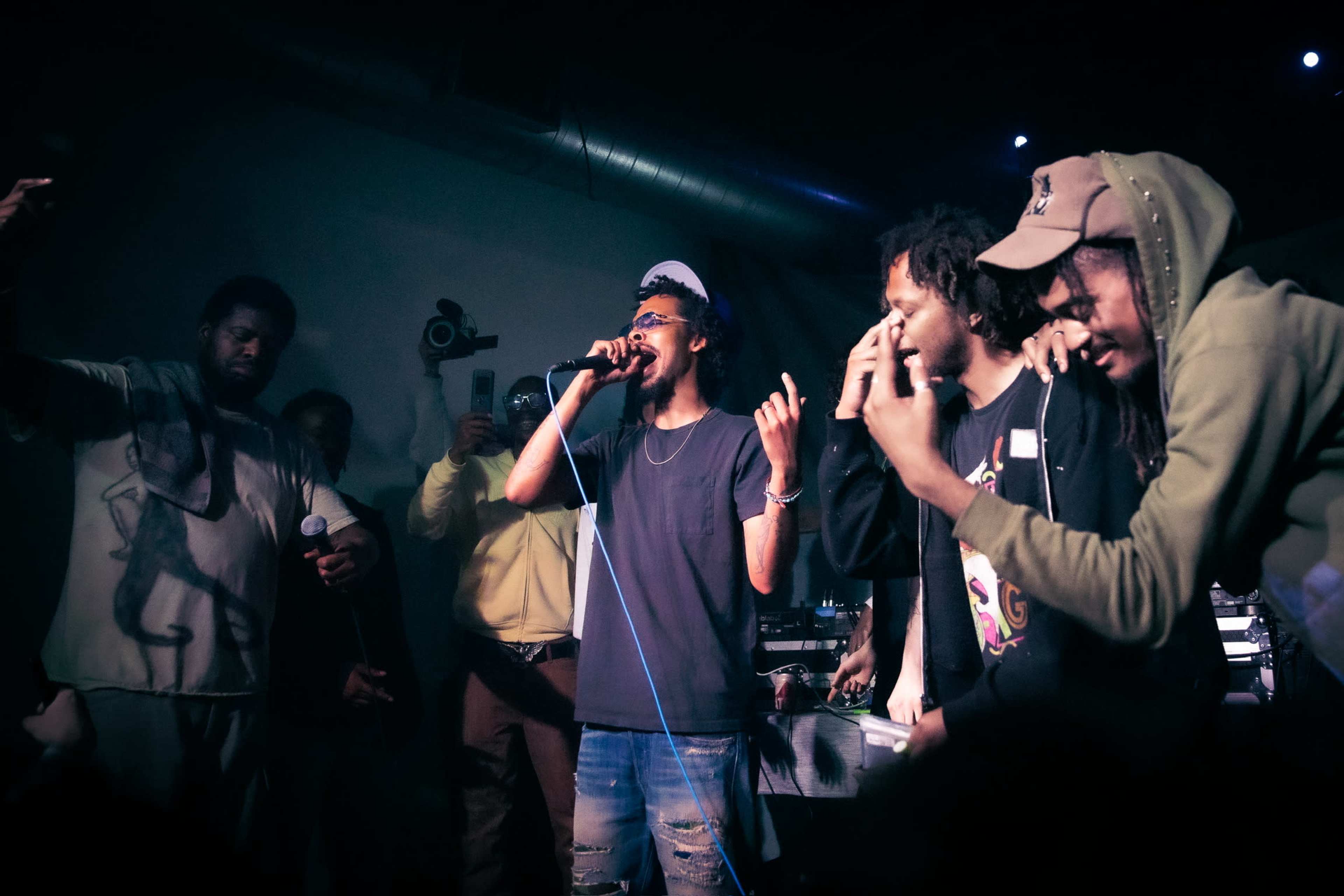 A group of performers stands on stage, with one artist singing into a microphone while others engage with the audience and each other.